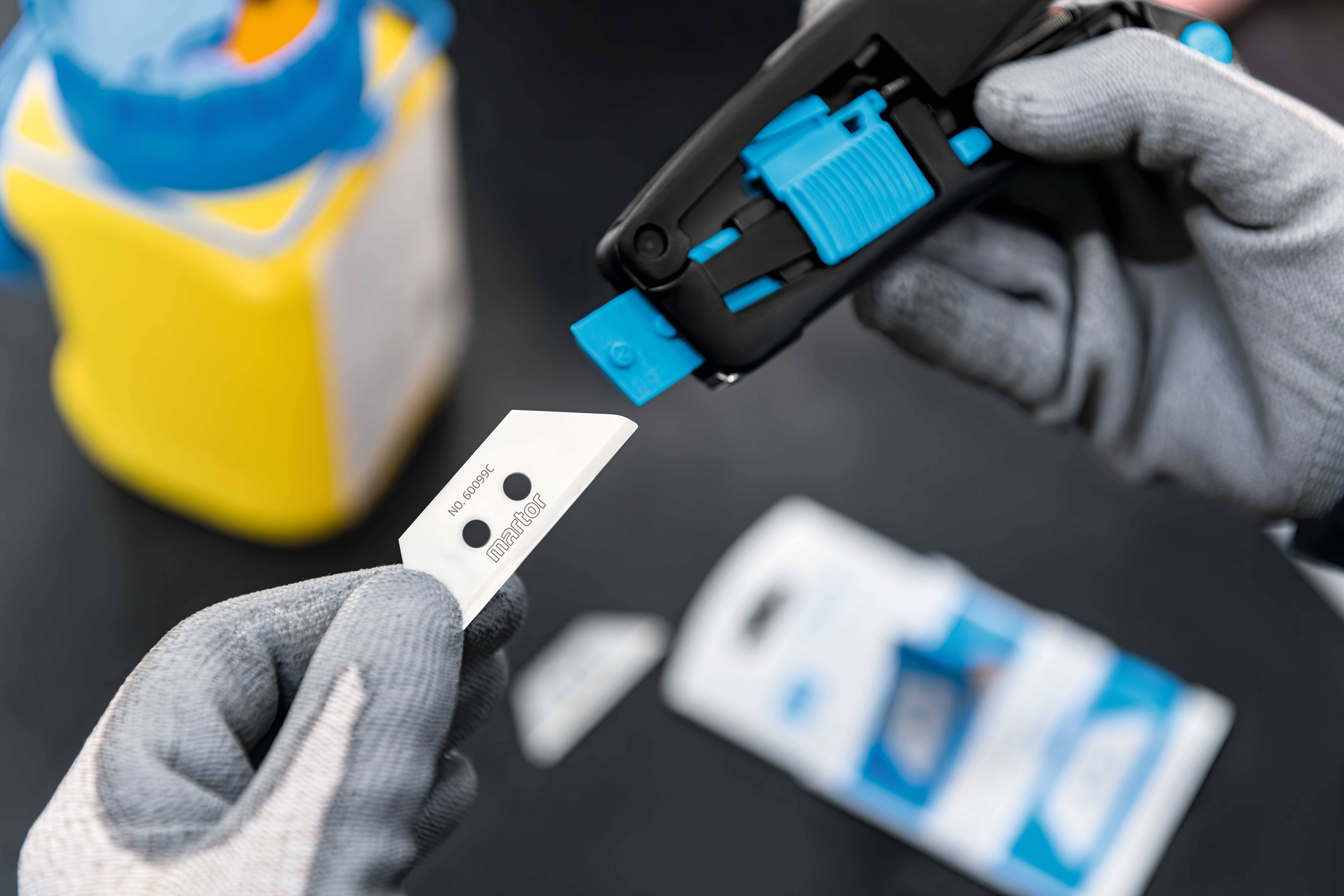 A hand pulls a blade from a black blade dispenser, with a blurred yellow object in the background. Topic: safe blade changing.