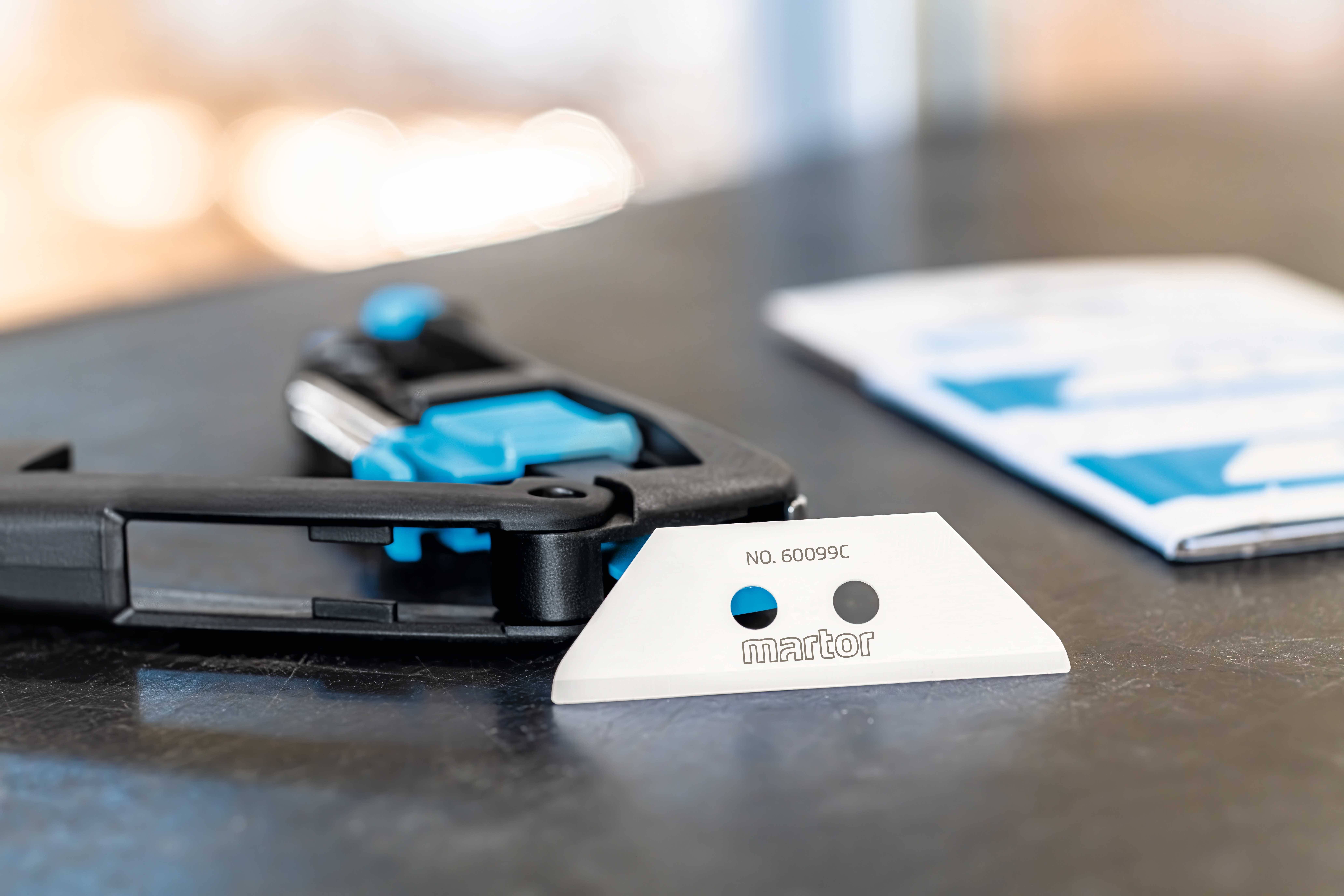 'Martor Safety Blade No. 60099C' on a table, with a blue safety knife in the background.