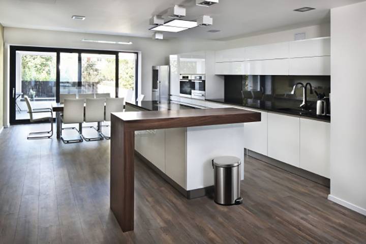 Modern kitchen with white cupboards, central wooden island and large windows overlooking the exterior. Dining area with white chairs.