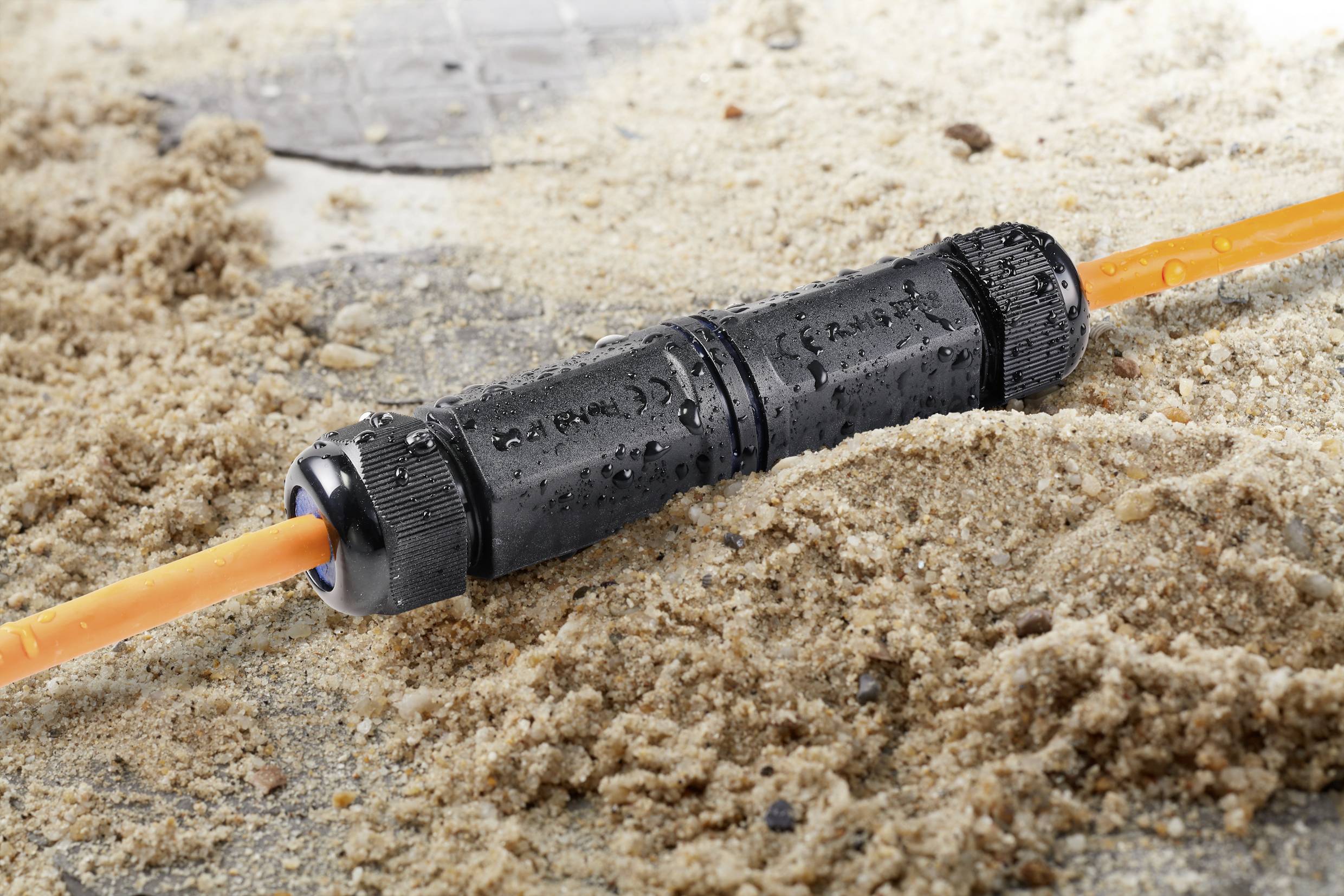 A waterproof cable connector on a sandy surface, with two orange cables connected to it.