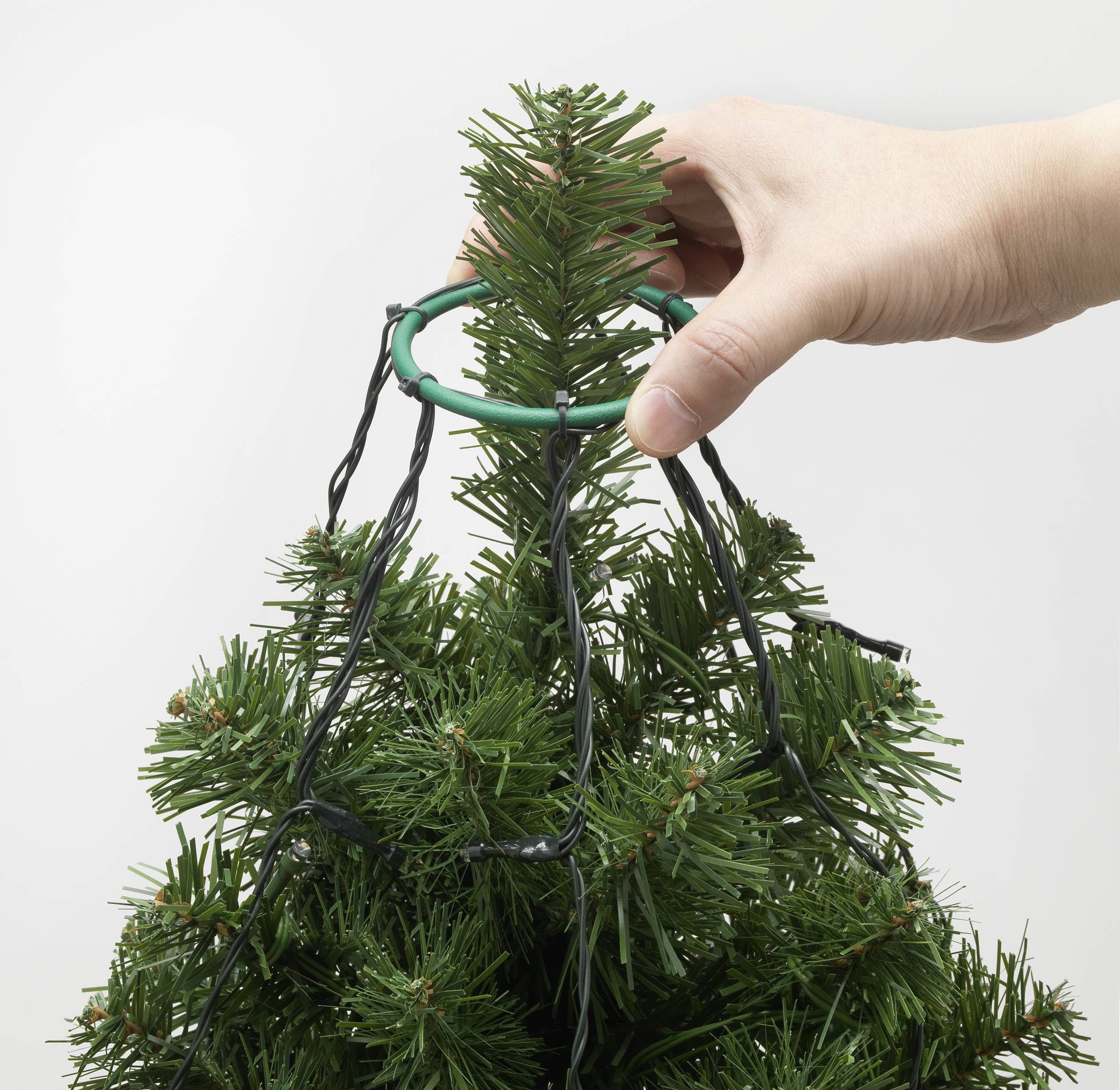 A hand places a green plastic clip at the top of an artificial Christmas tree to secure lights to it.