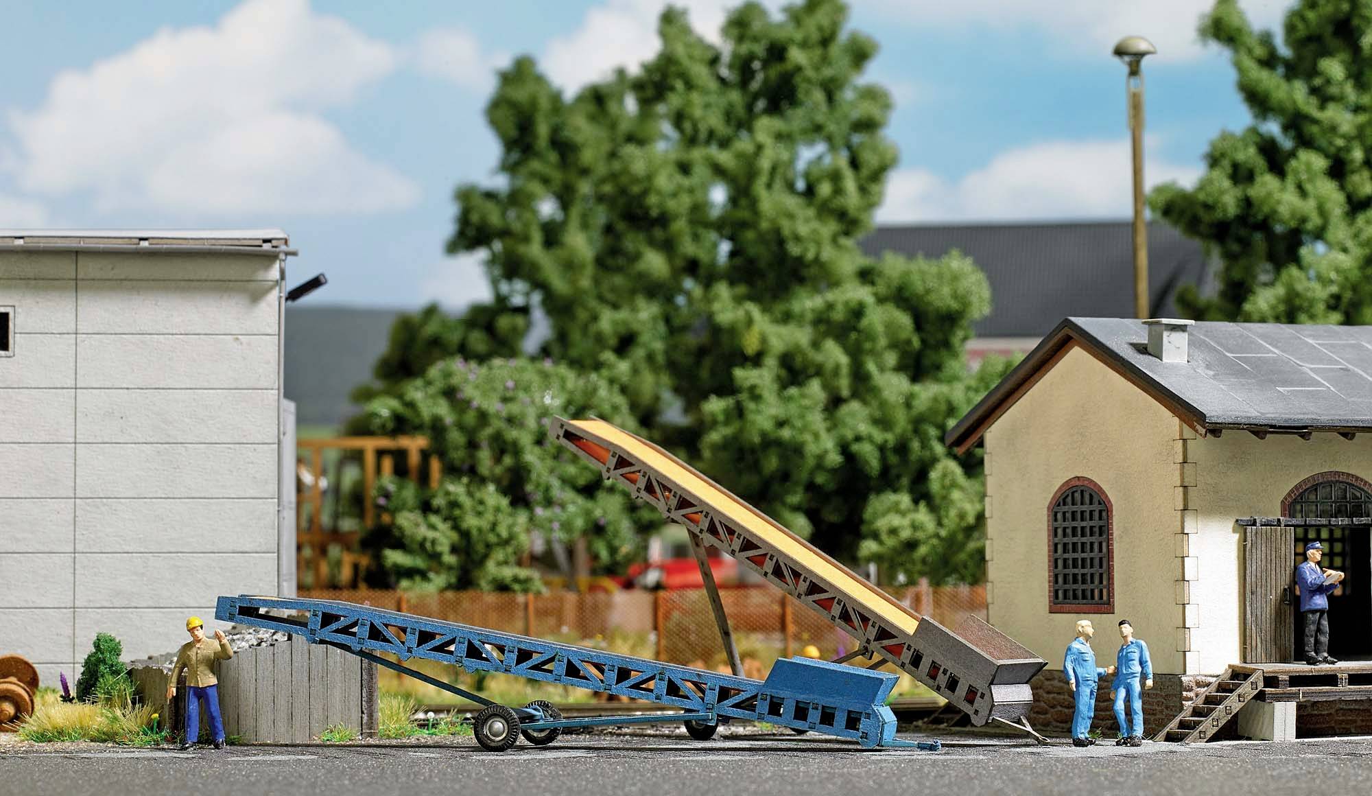 A miniature model shows people working beside a conveyor system between two buildings on an industrial site.