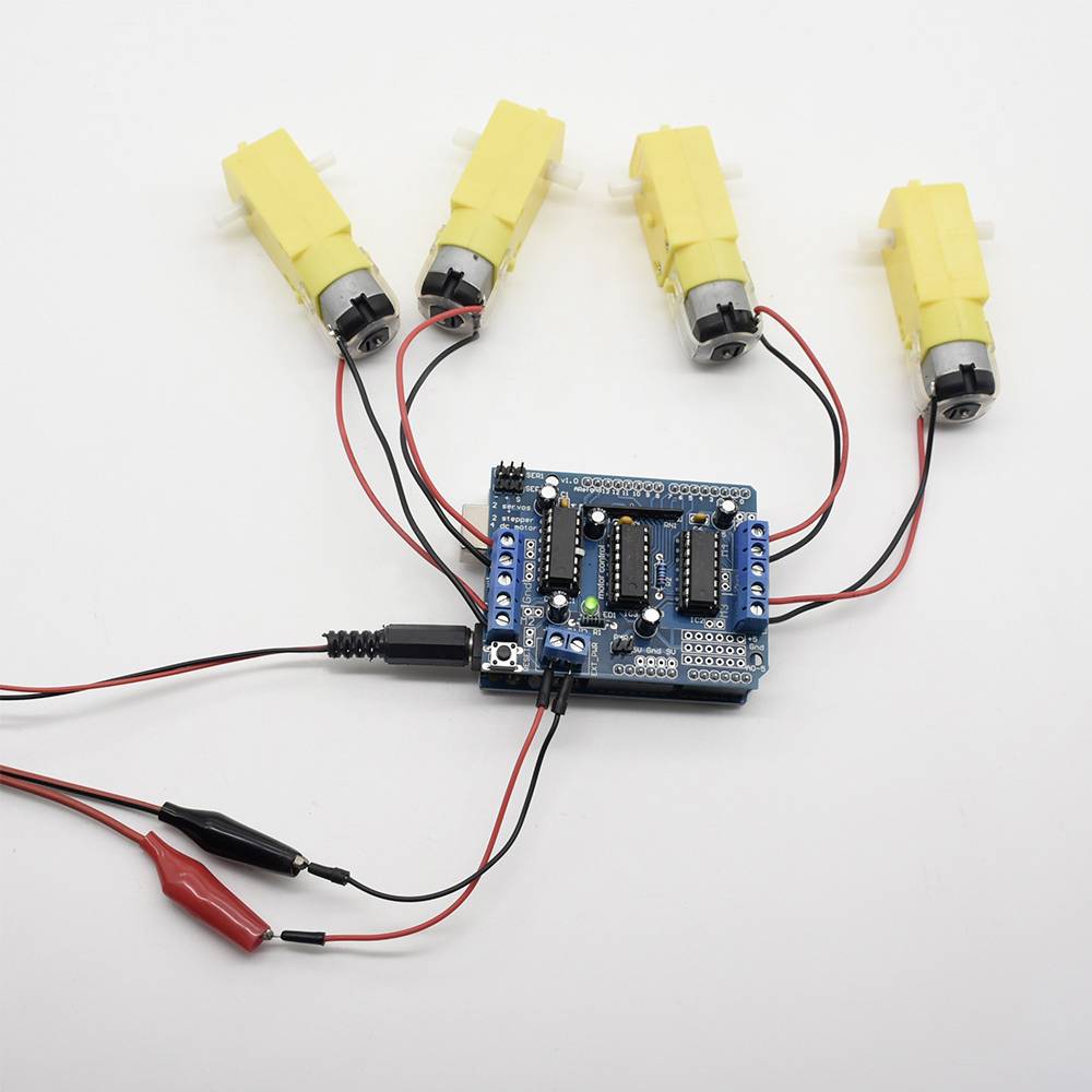 A blue circuit board is connected to four yellow motors and red and black cables. The circuit board is illuminated by green LEDs.