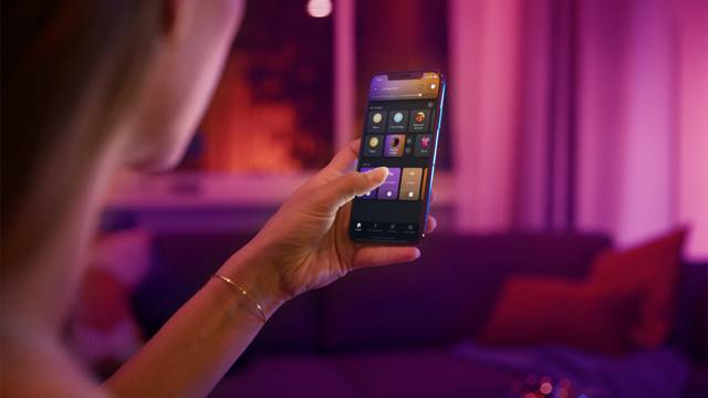 A person is operating a smartphone to control the lighting in a living room. The room is illuminated in warm colours.
