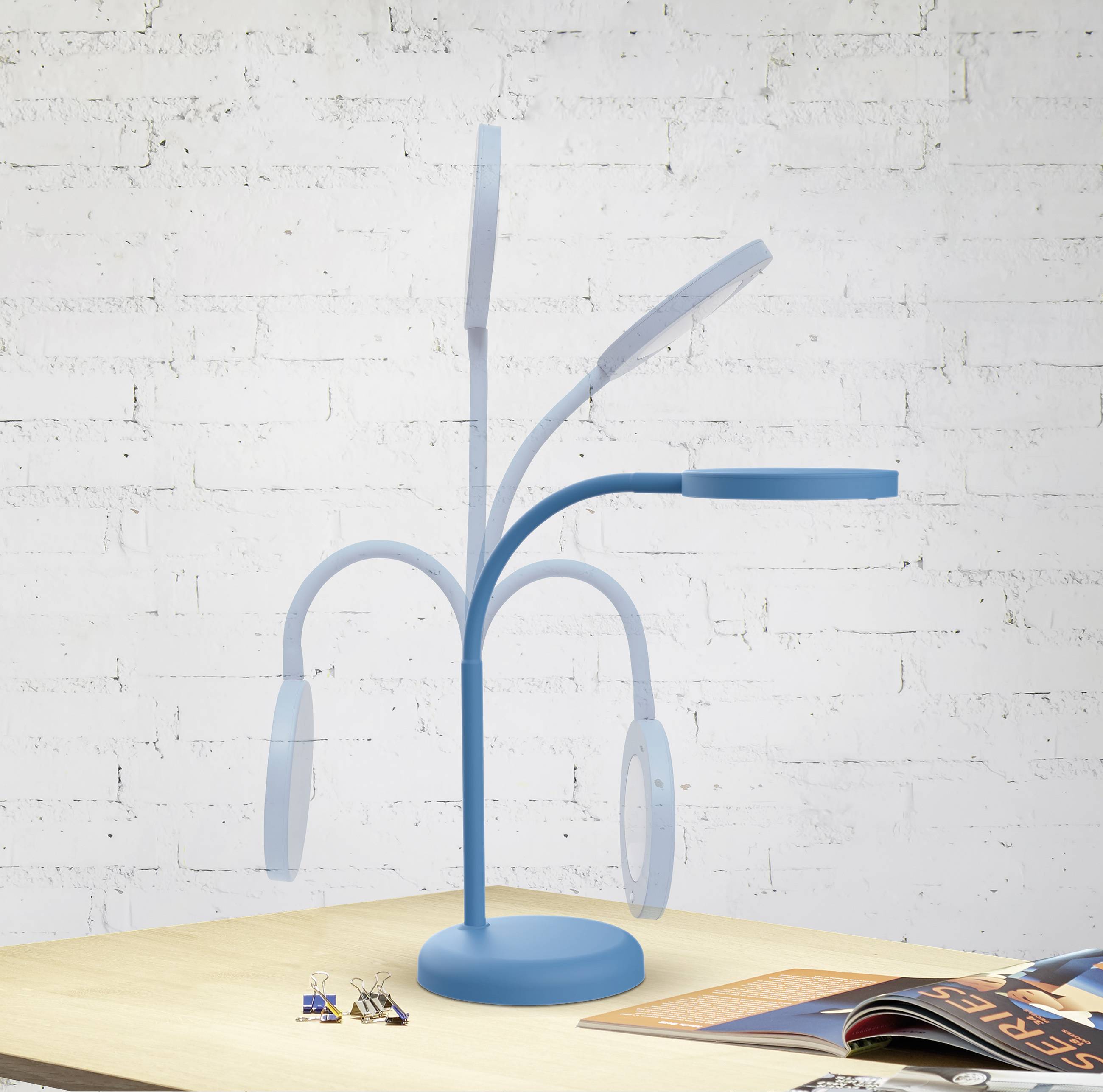 A blue-painted desk lamp with flexible arms sits on a wooden desk. Keys are located on the left, an open magazine on the right.