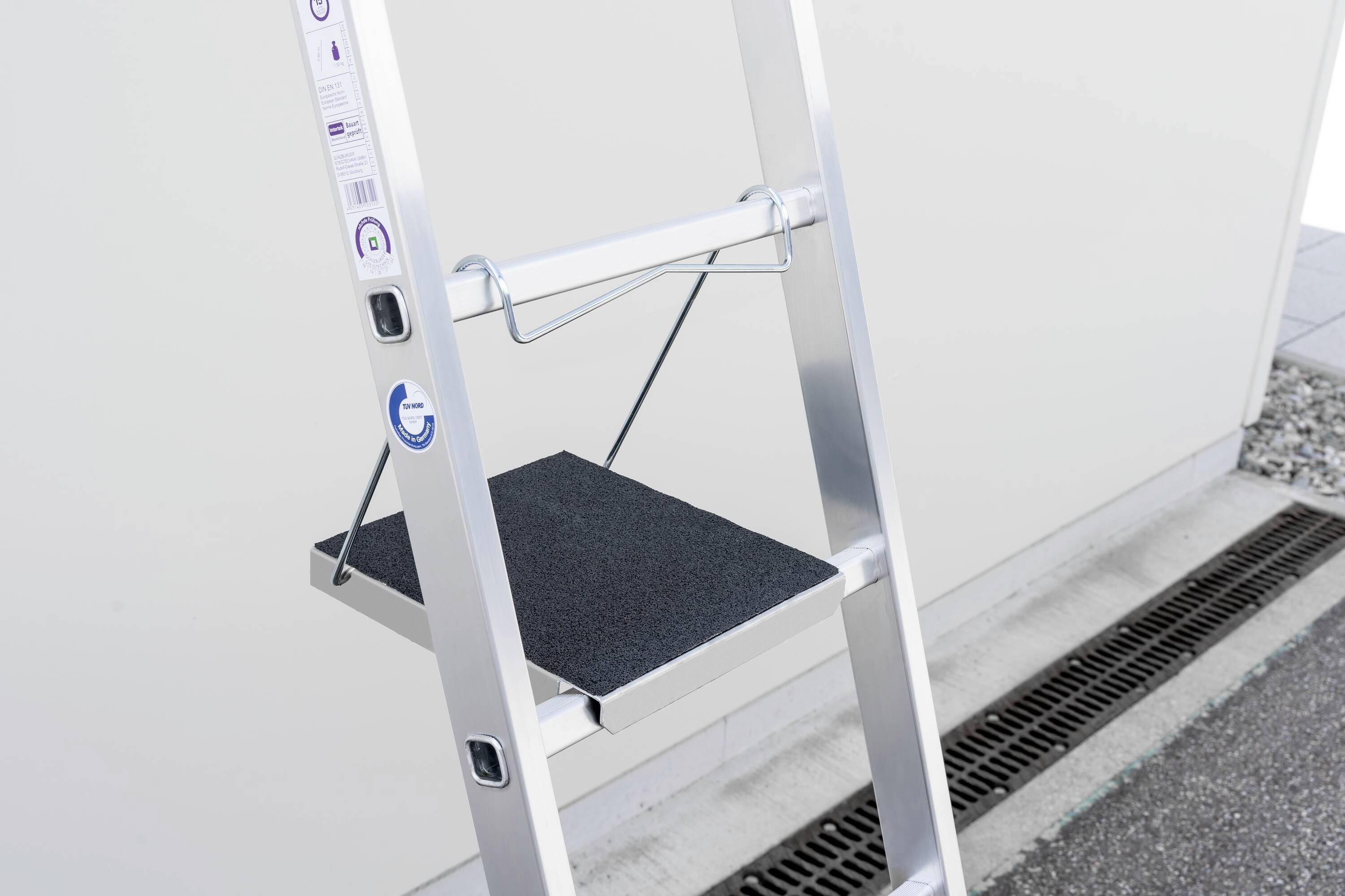 Aluminium ladder with extendable black platform against a grey wall. Suitable for versatile use.