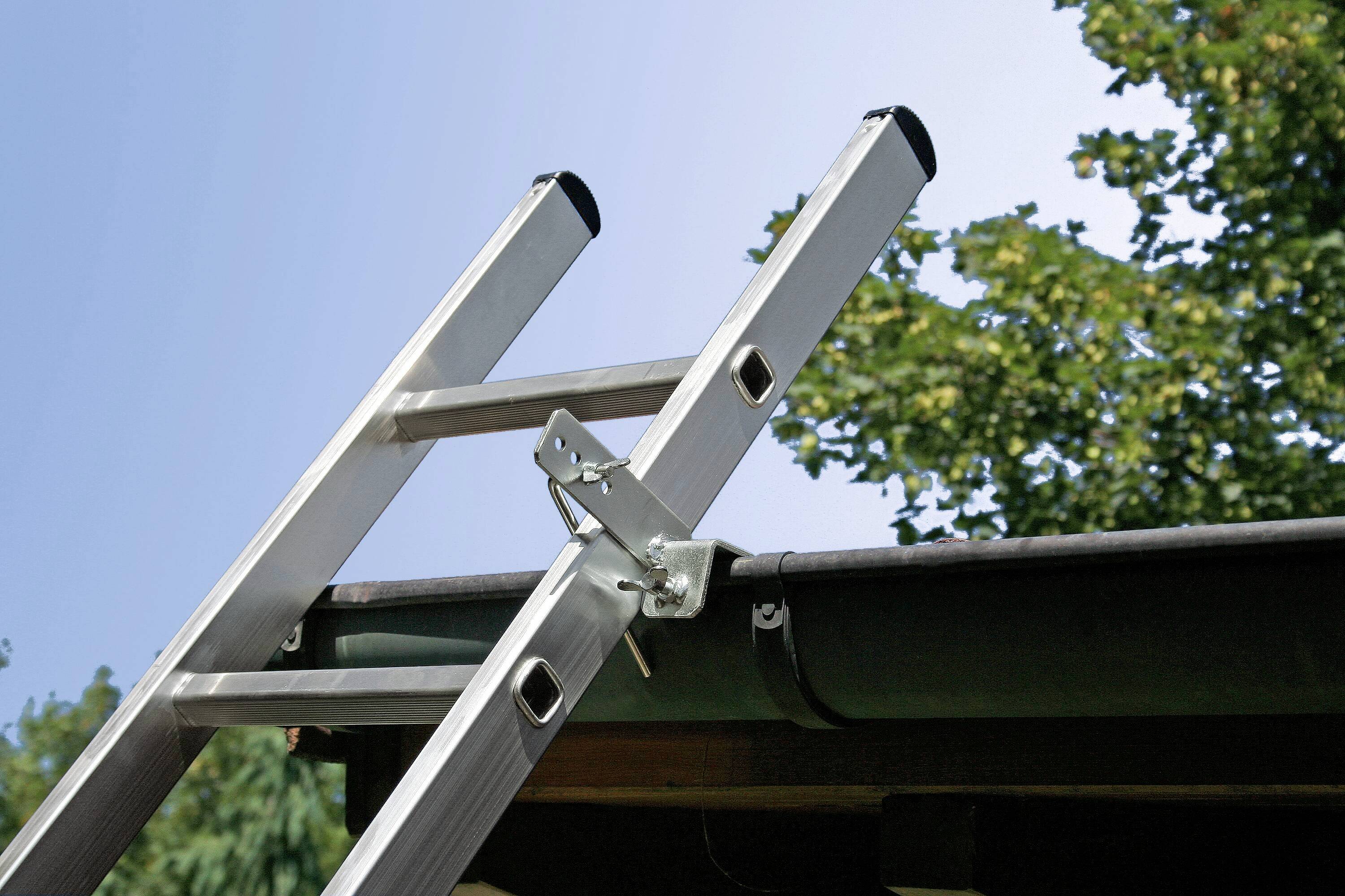A ladder is leaning against a gutter on a roof beneath a clear sky and green trees, indicating work at height.