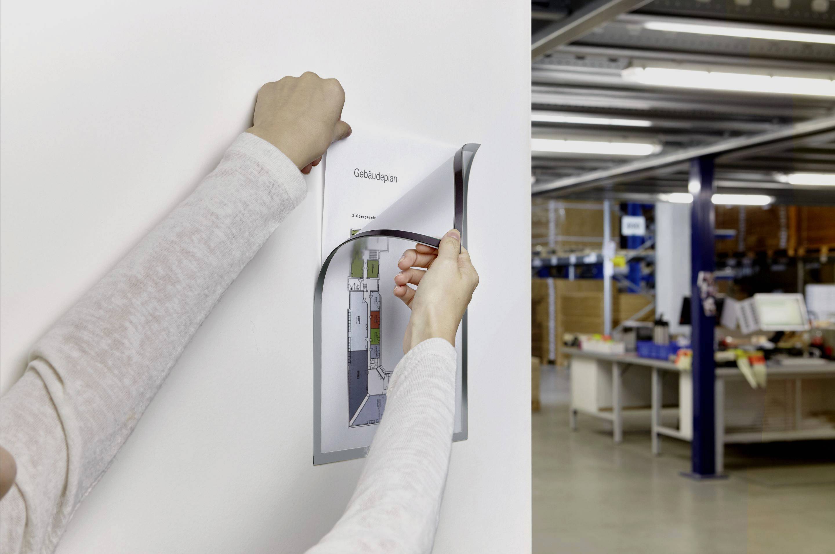 A person is hanging an information sheet titled 'Building Plan' on a wall. Office furniture can be seen in the background.