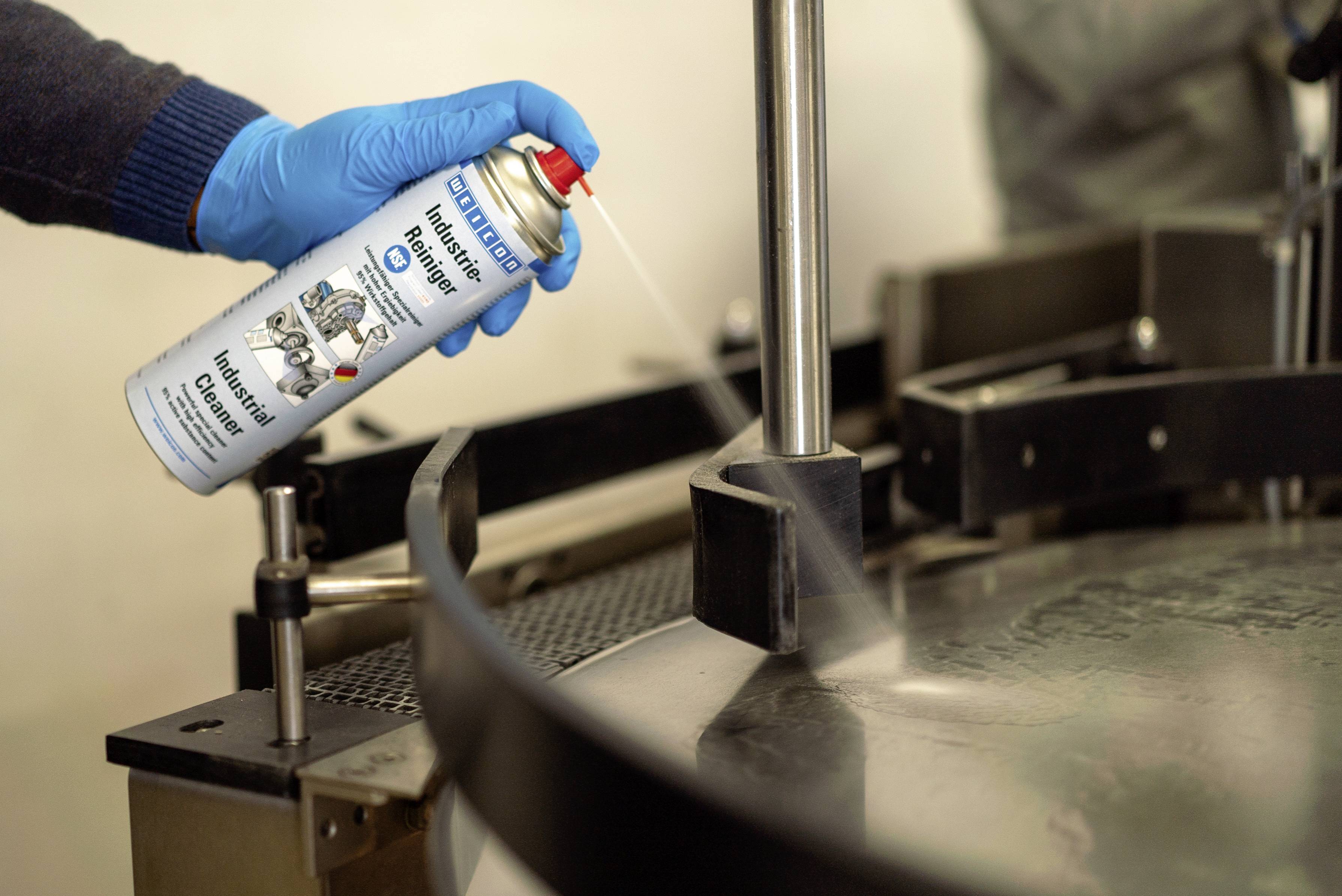 A person wearing blue gloves is spraying lubricant spray onto a metal device in an industrial setting.
