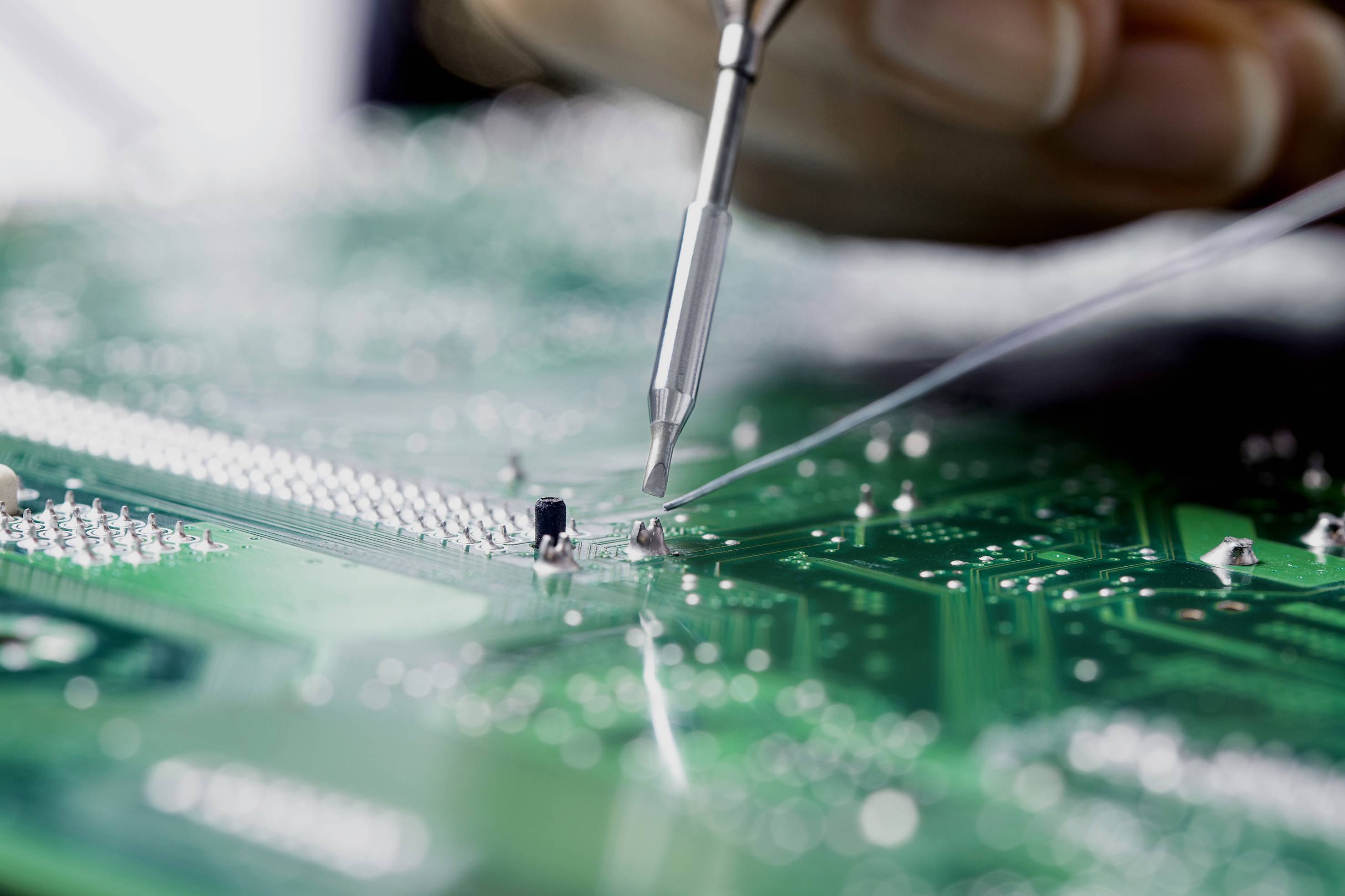 Close-up of a soldering iron working on a green circuit board, focusing on detailed circuitry and components.