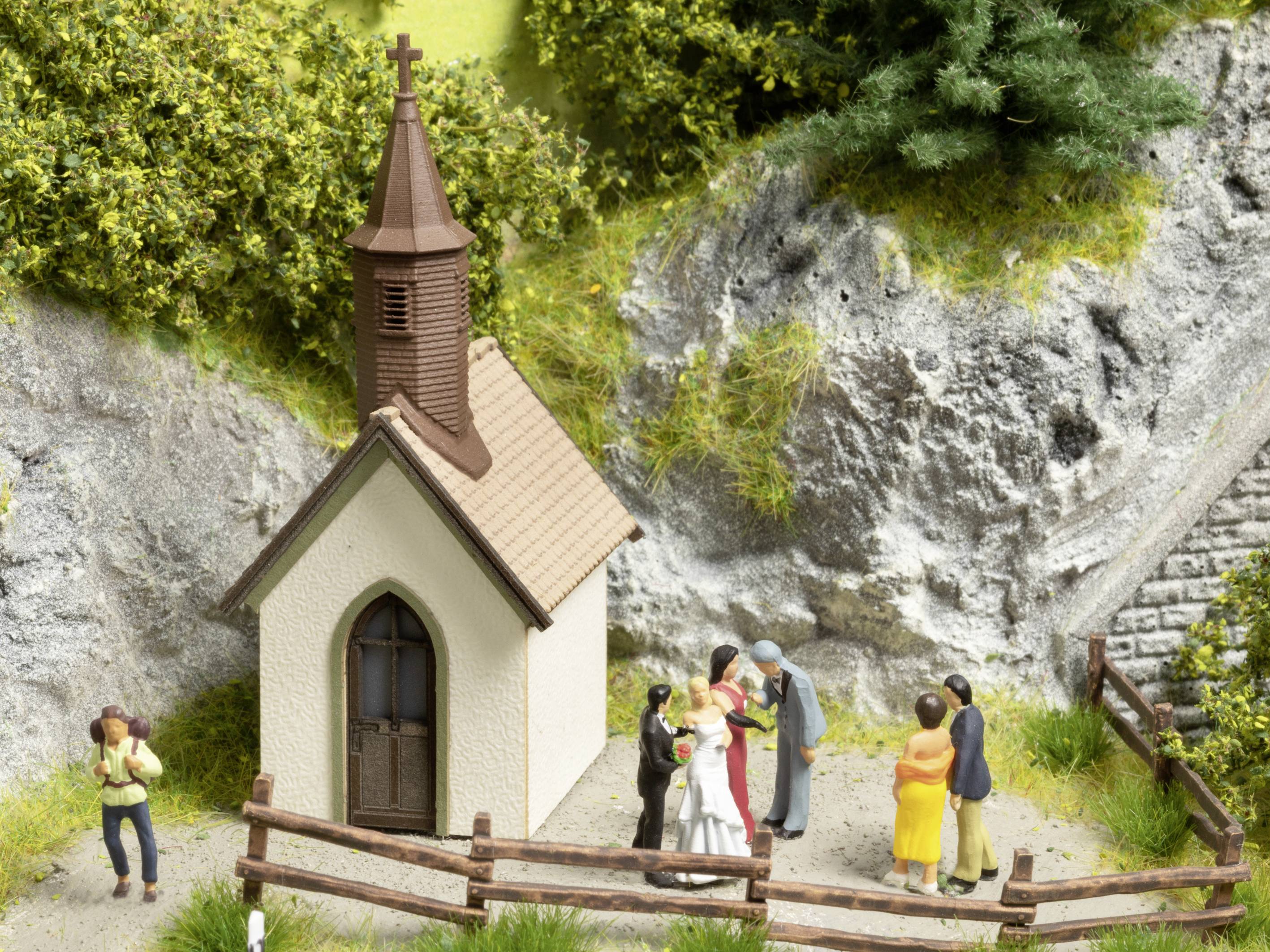 A small chapel on a hill. A wedding with a bride and groom, guests, and a photographer is taking place in front of it.
