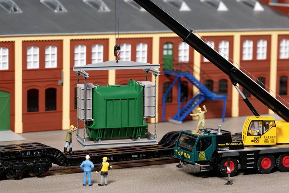 Model of an industrial scene: A crane is lifting a green transformer onto a goods wagon in front of a building complex. Several figures are observing.