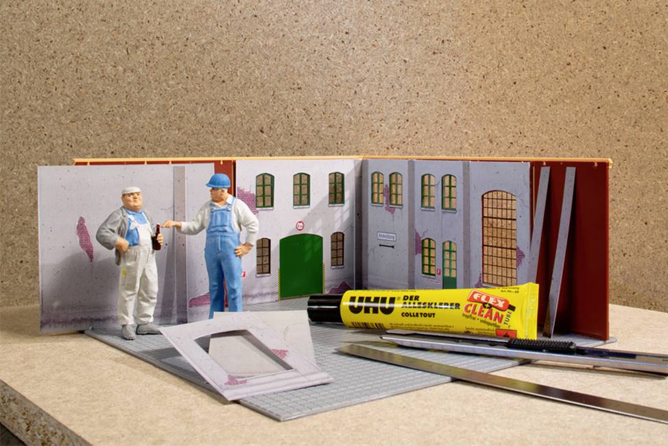 Two miniature figures stand in a model-building kit. Around them, tools and glue are scattered on a table. In the background, a section of a building can be seen.