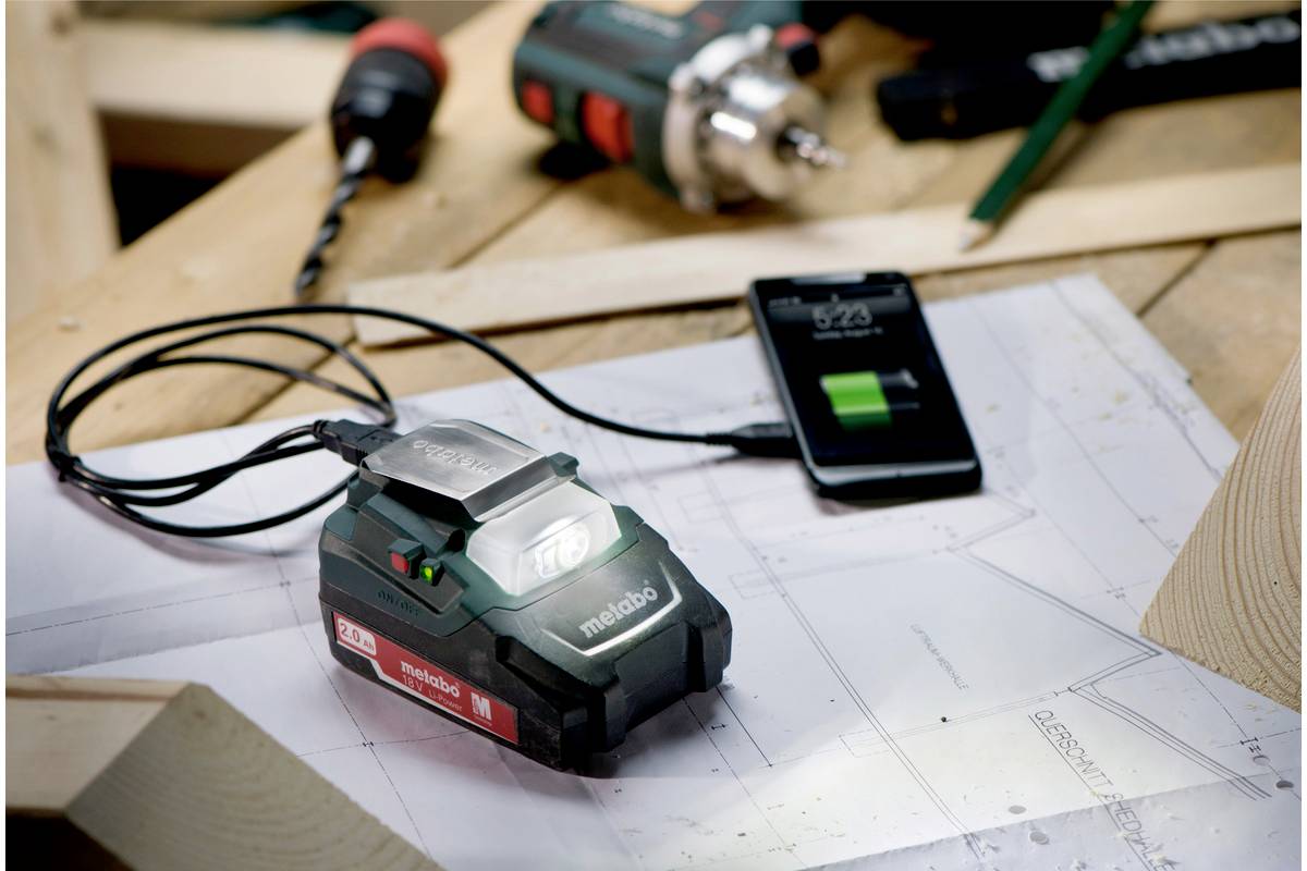 A charging station for electric tool batteries on a wooden table, connected to a smartphone, displays the charging process. Blueprints and tools are lying in the background.