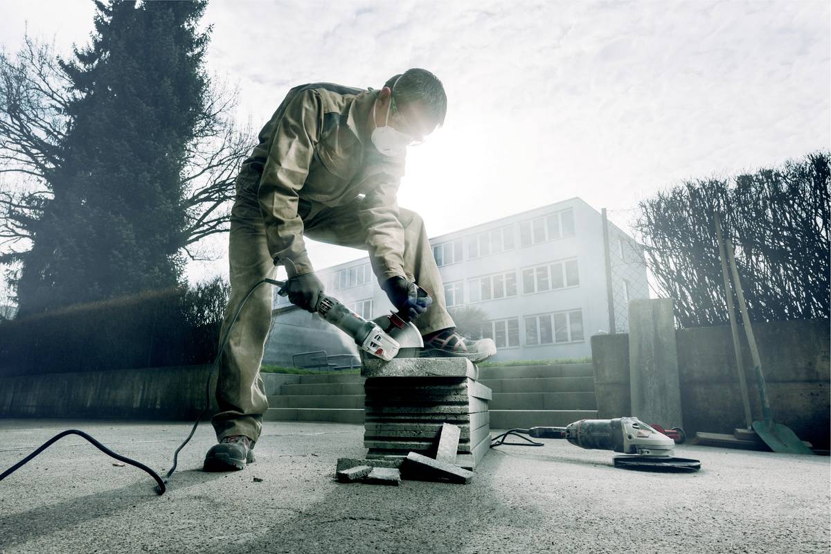 A man wearing protective clothing is cutting through stacks of floor tiles with an angle grinder on an outdoor terrace.