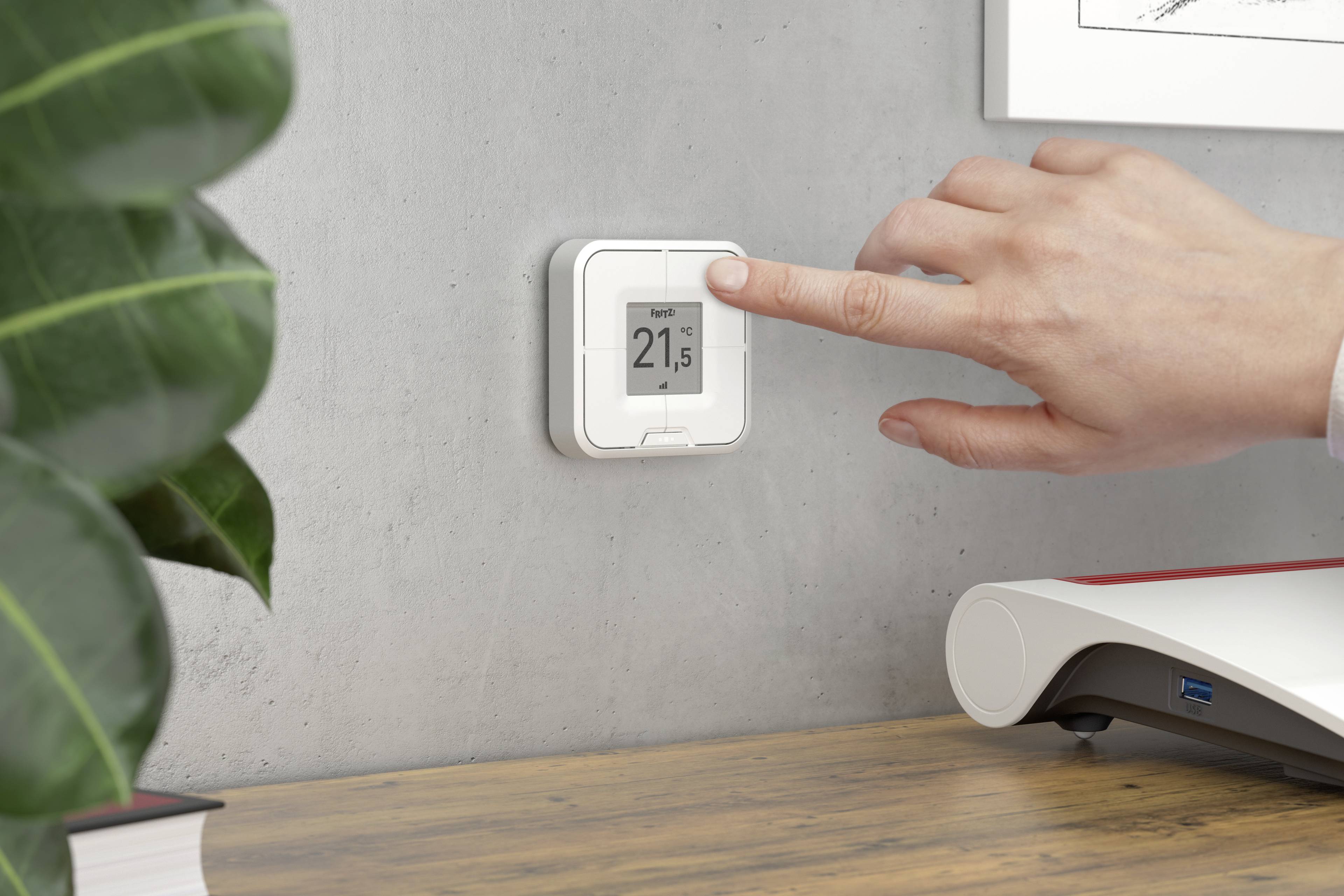 A hand adjusts the digital thermostat on a concrete wall to 21.5 degrees. Beside it is a plant and a desk.