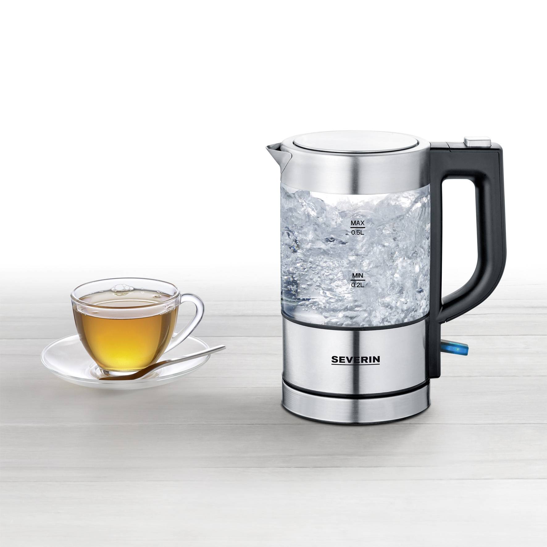 A kettle with boiling water next to a cup of tea on a table. The kettle shows maximum and minimum water level lines.