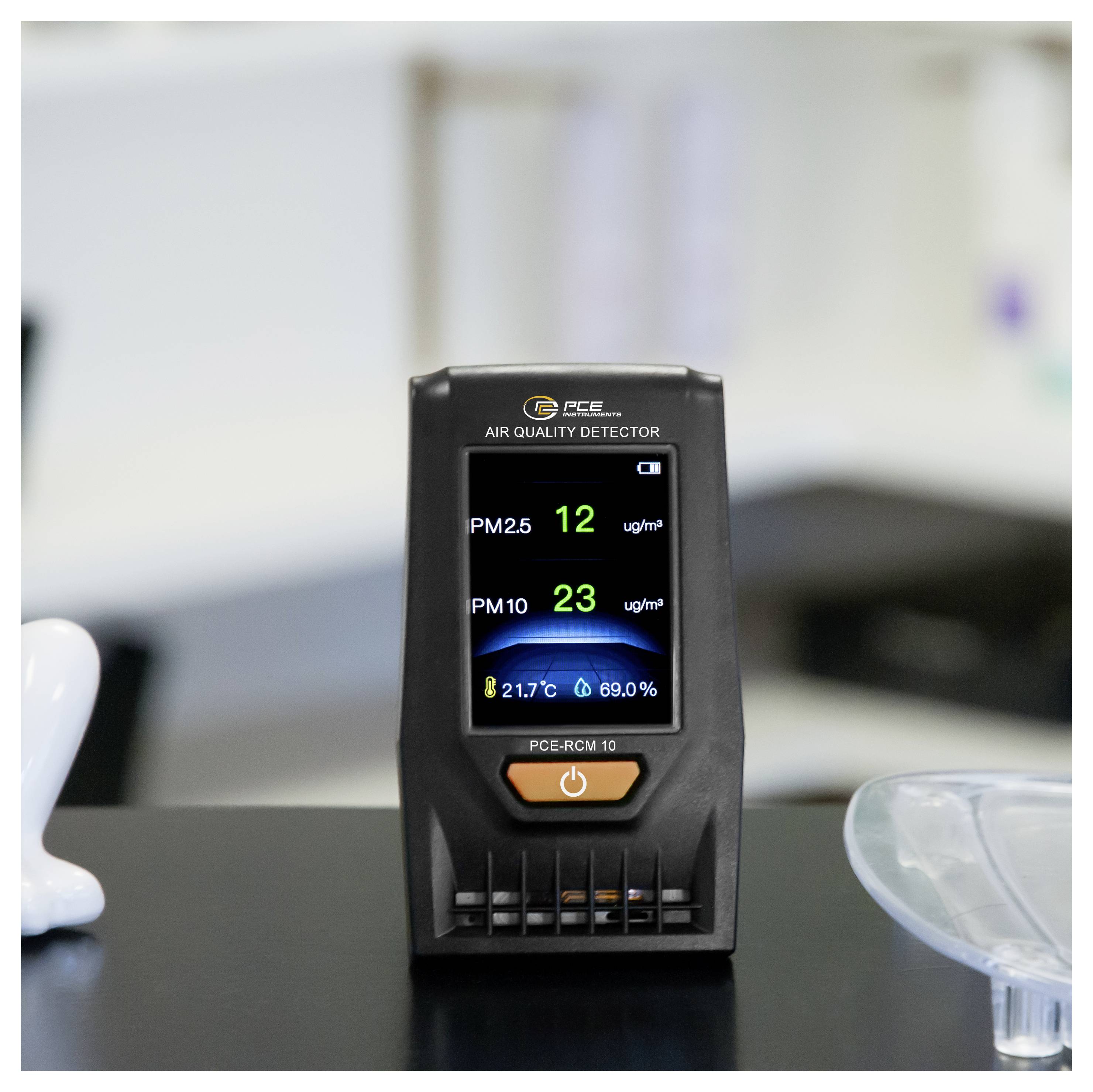 Portable air quality detector on a desk showing PM2.5 12 ug/m³, PM10 23 ug/m³, 21.7°C, and 69.0% humidity.