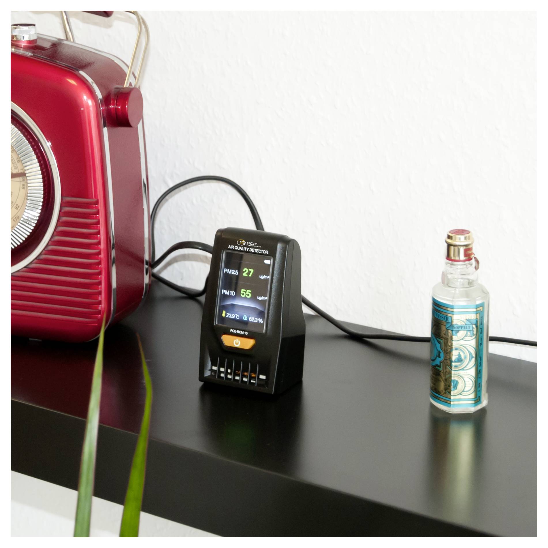 A small air quality monitor on a black shelf reads PM2.5 27 and PM10 55, beside a red radio and a vape device.