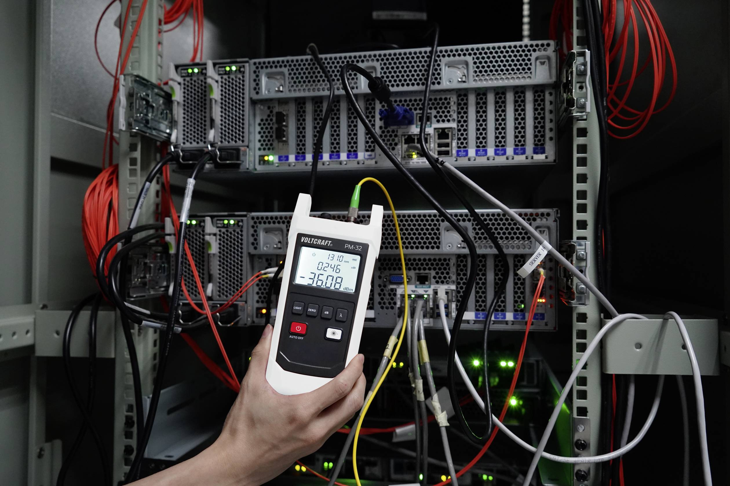 A hand holds a measuring device displaying the temperature in a server room. Several servers with cables are visible in the background.