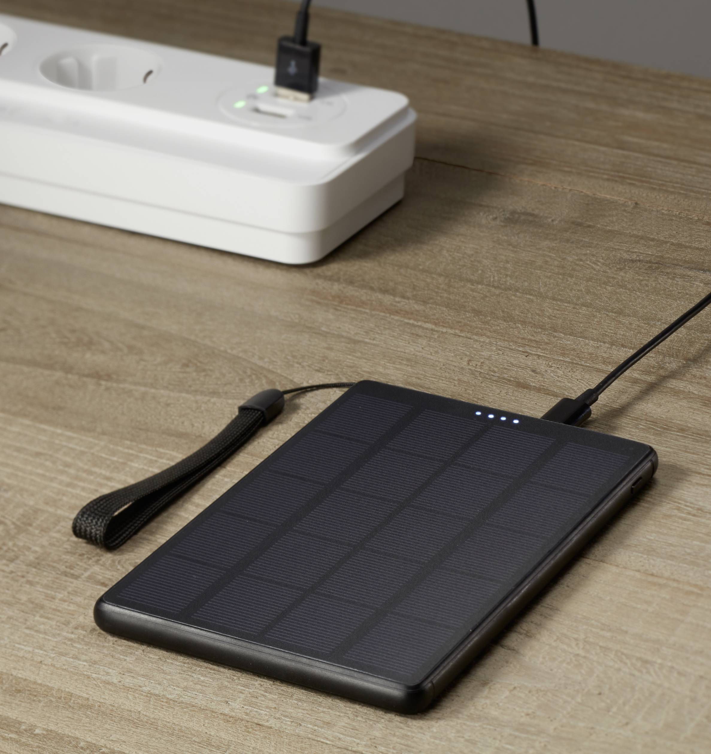 Solar-powered power bank with charging indicator, lying on a wooden table, connected via a cable to a power strip.