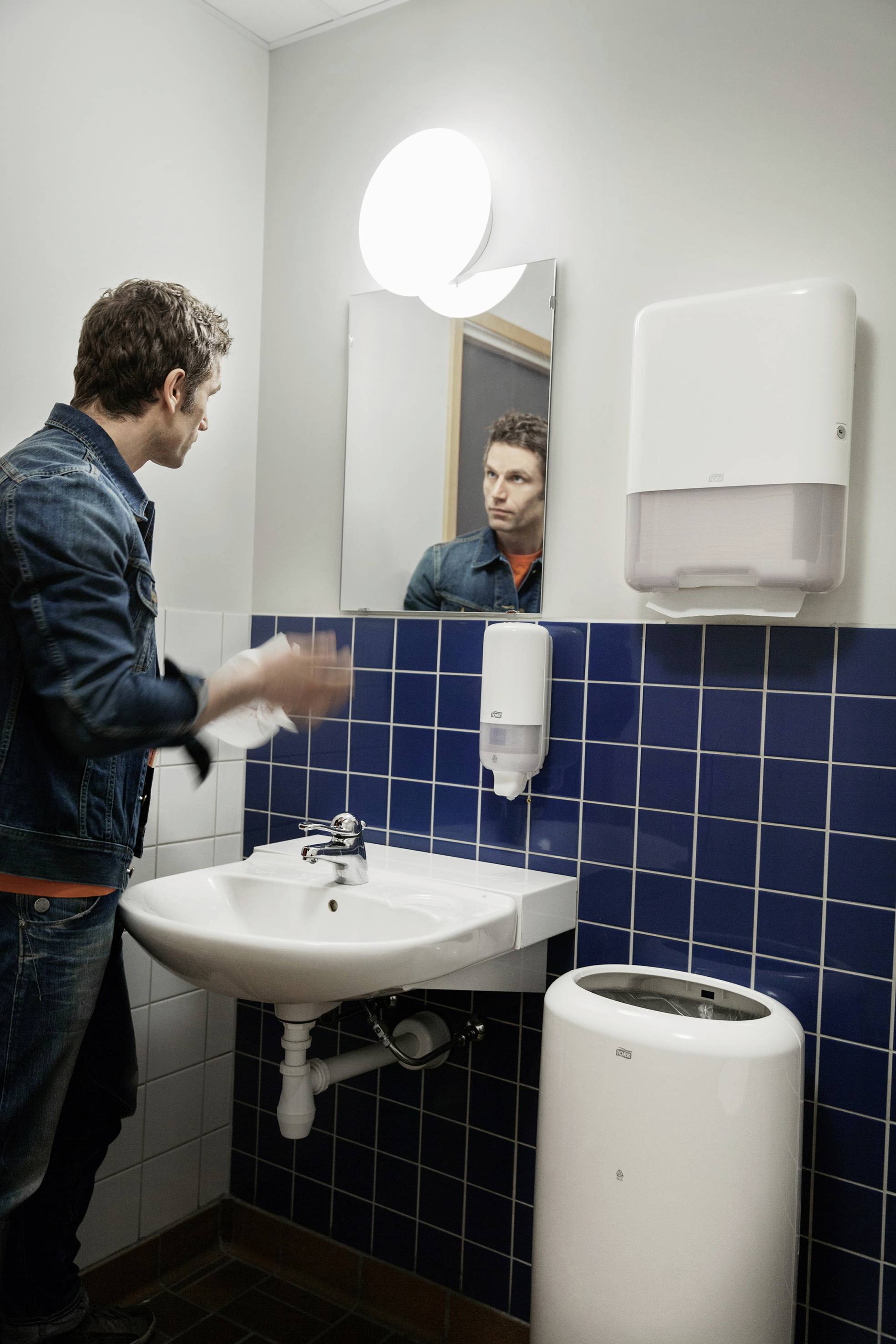 A man is washing his hands in a bathroom with blue tiles. A mirror, soap dispenser, and paper towel dispenser are visible.