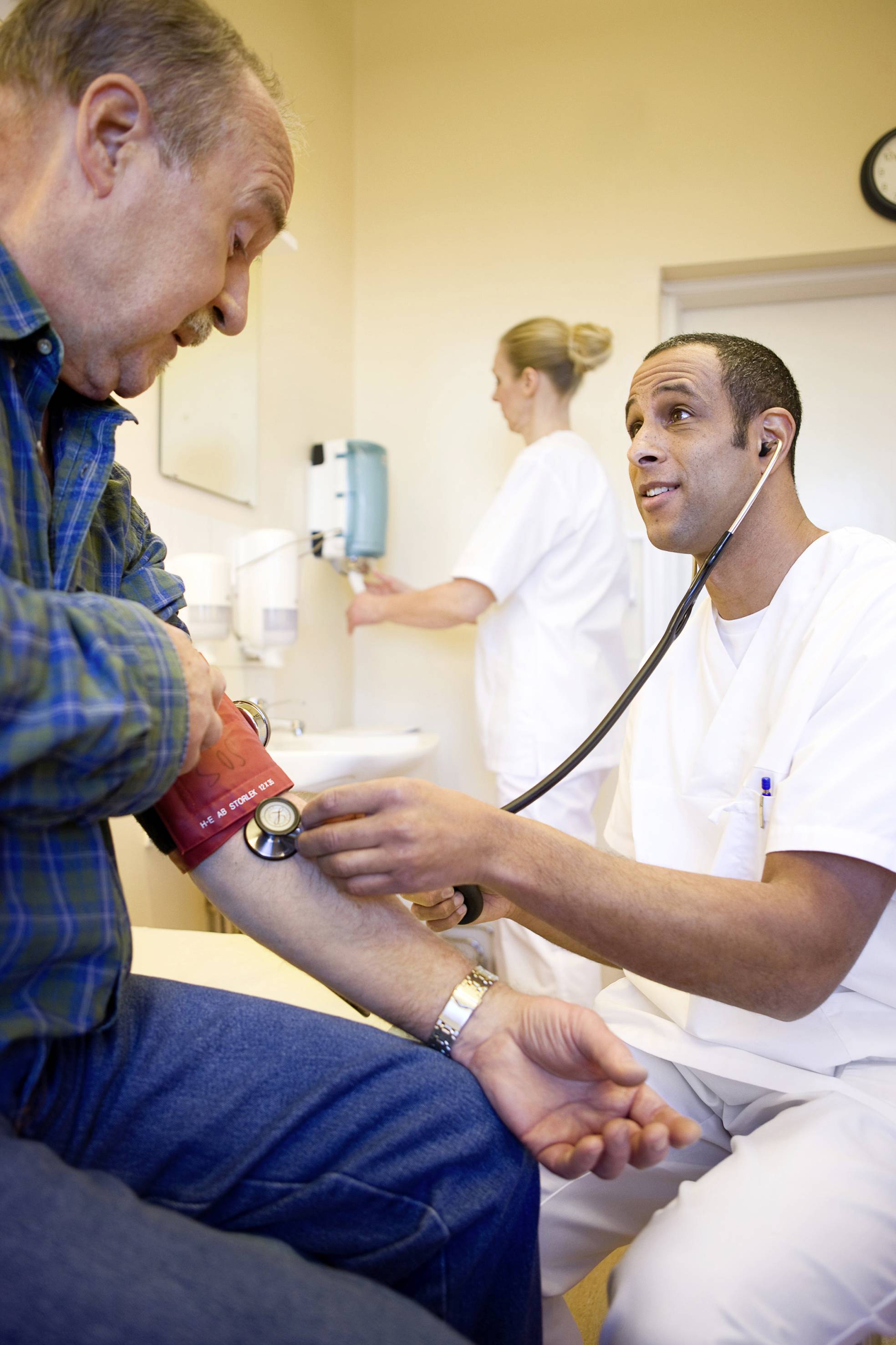 A doctor measures the blood pressure of an elderly man in a medical practice, while a nurse works in the background.