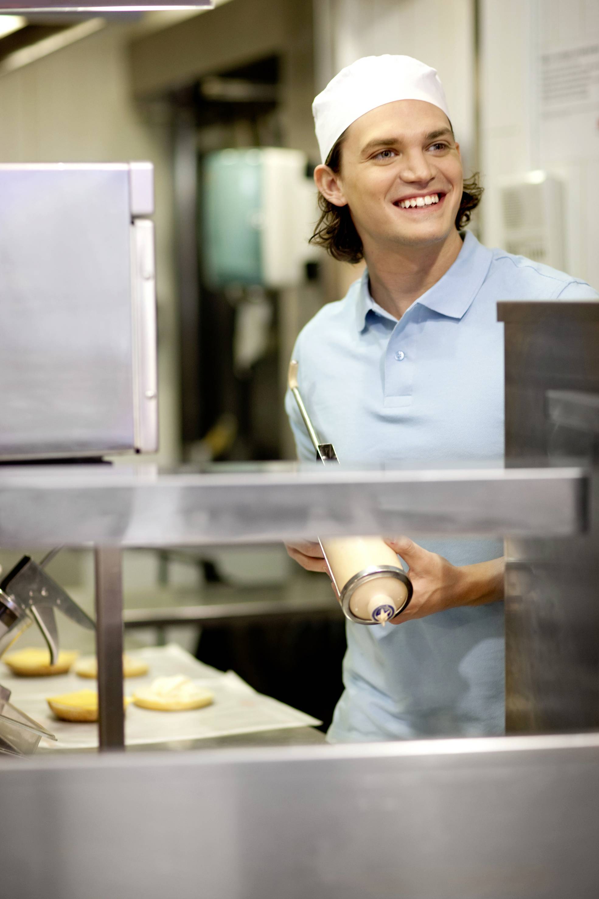 A young man in chef's attire stands in a kitchen, holding a sauce bottle, smiling while preparing sandwiches.