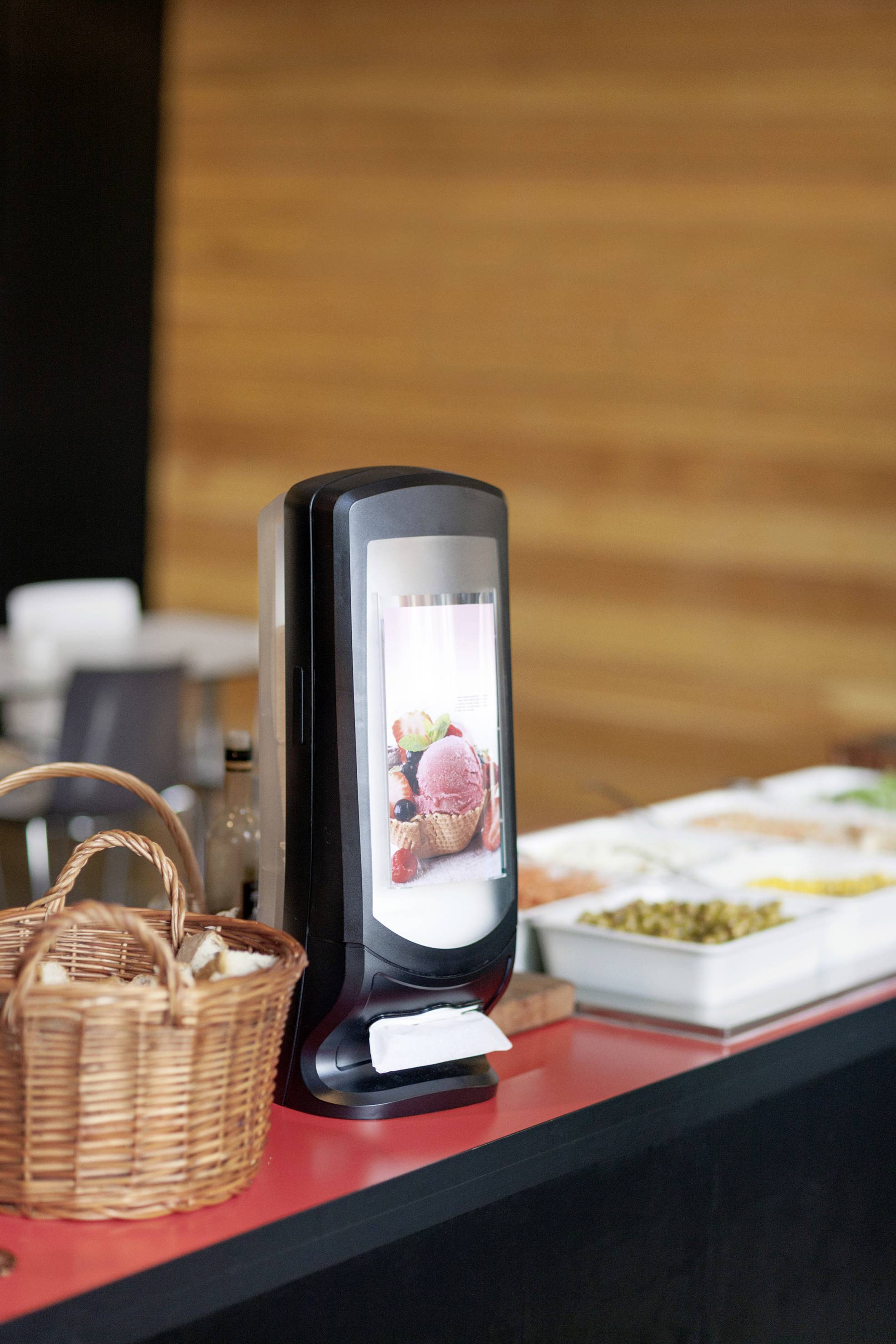 Ice cream dispenser on a red counter with serviettes, beside a woven basket and various condiment containers.
