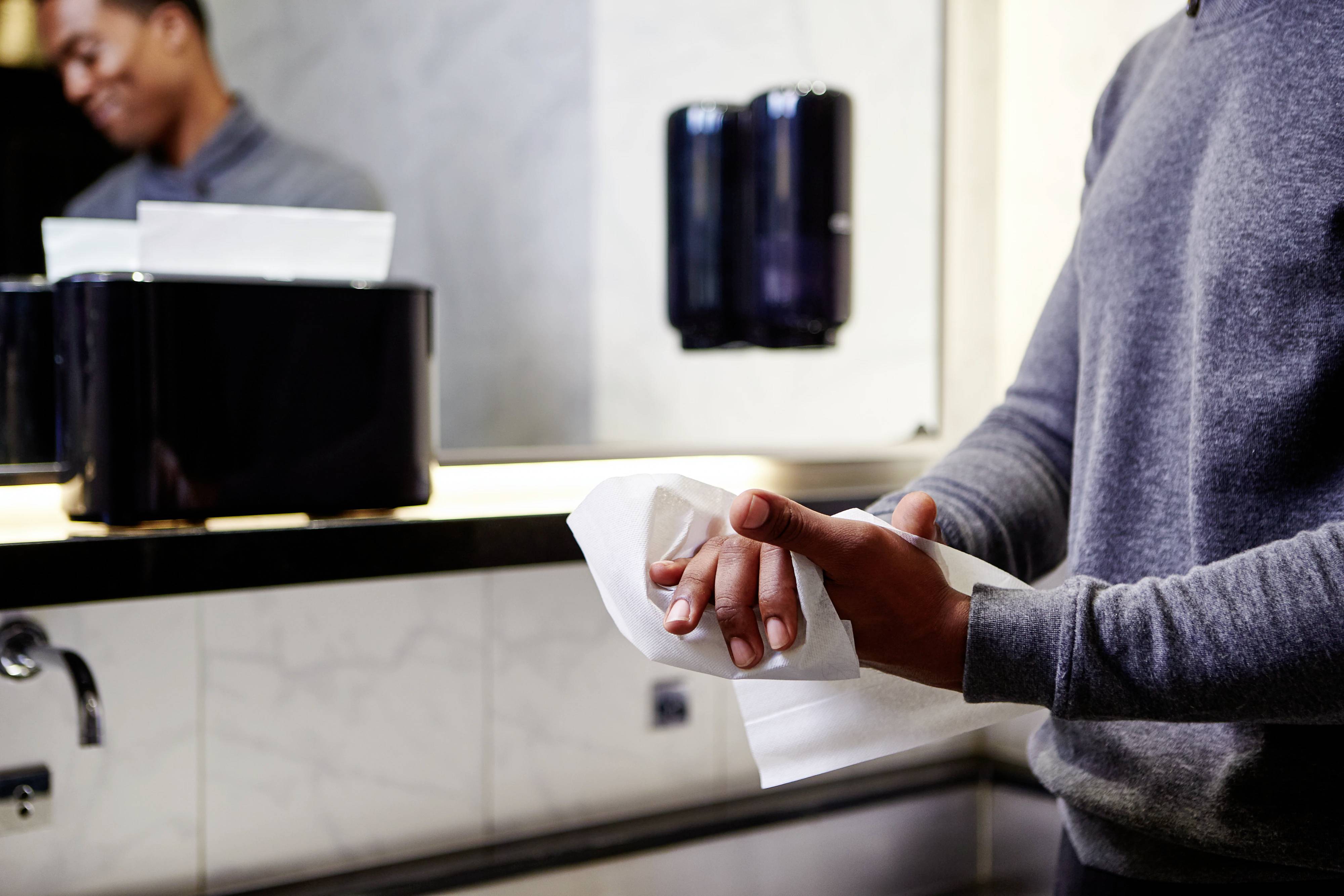 A person is drying their hands with a paper towel in a bathroom. A mirror and soap or towel dispenser are visible in the background.