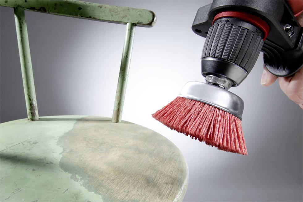 A person is sanding the seat of an old metal chair with a red rotary wire brush in order to renovate it.