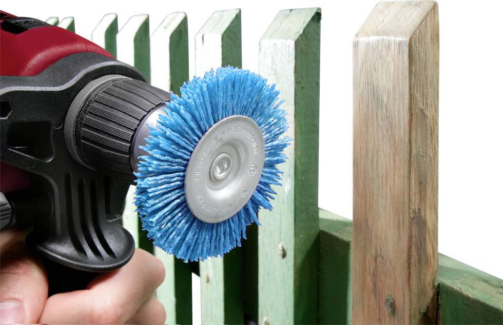 A drill with a blue brush attachment is being used to clean or sand the surface of a green wooden fence.