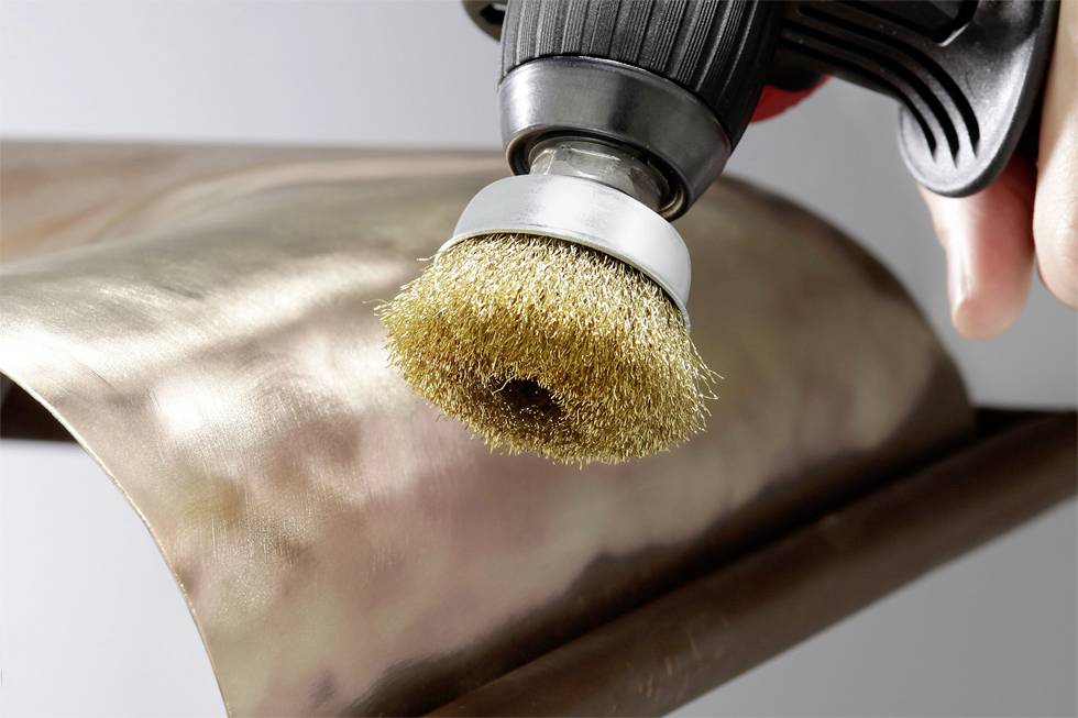 Electric drill with wire brush attachment polishes copper metal surface, focus on brush head.