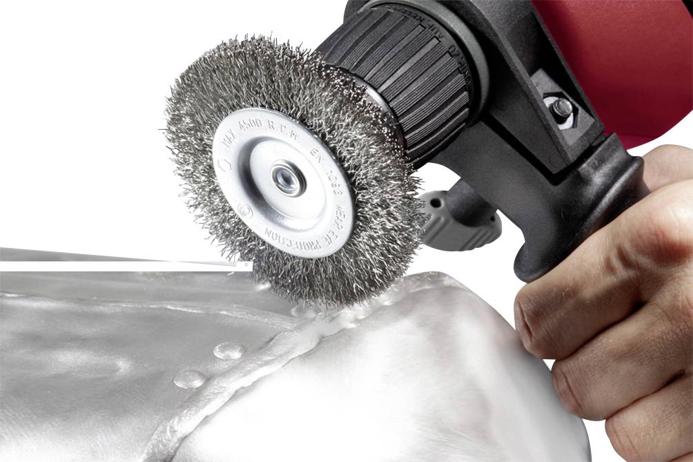 A hand is using an electric wire brush to grind or clean a metal surface, possibly a weld seam.