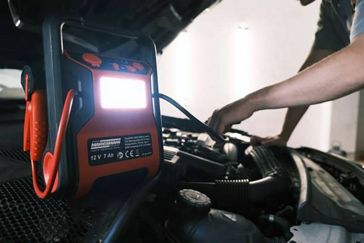 A person is using a portable jump starter to start a car with the bonnet open.