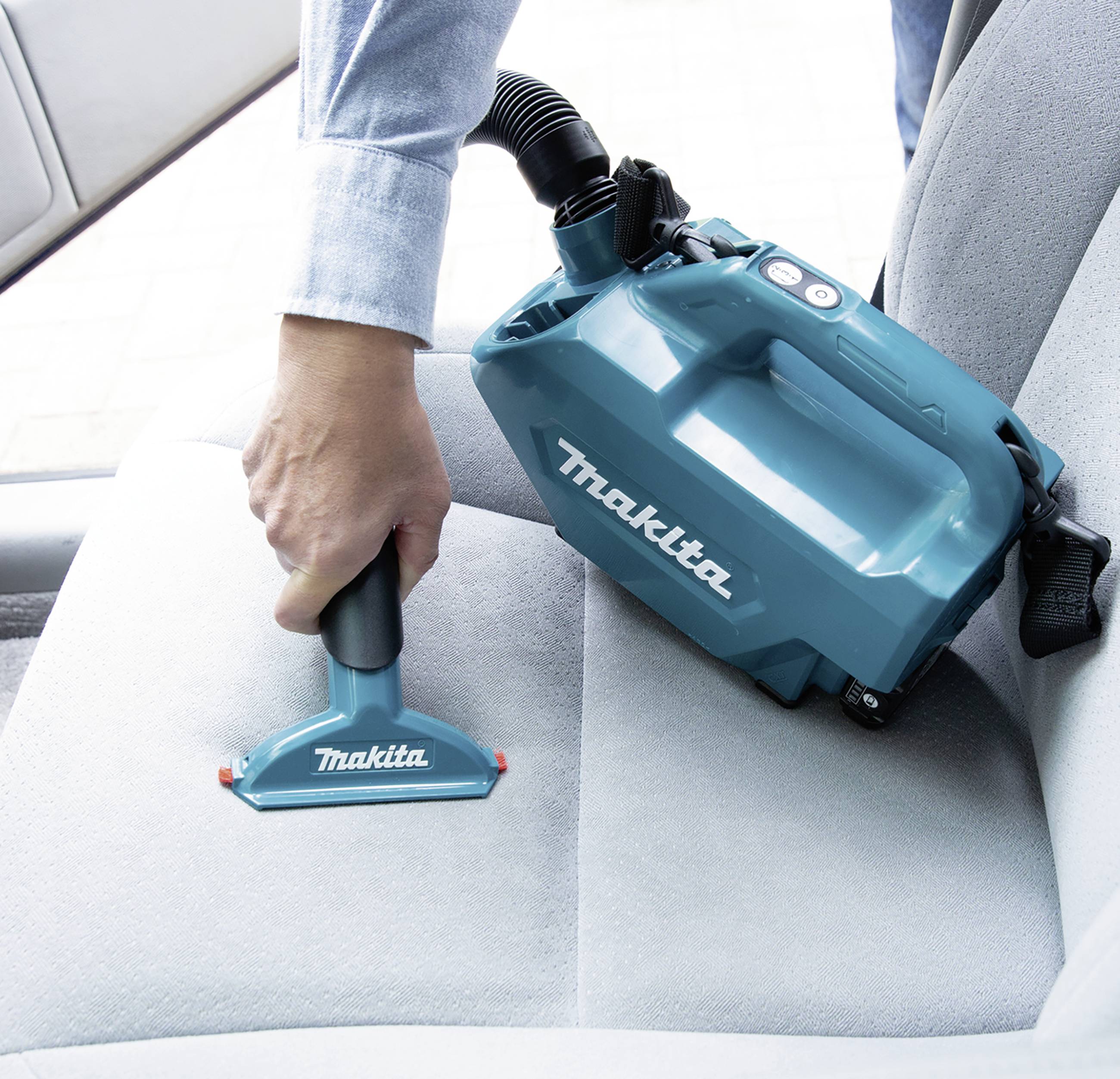 A person is holding a blue cordless Makita handheld vacuum and cleaning a car seat.