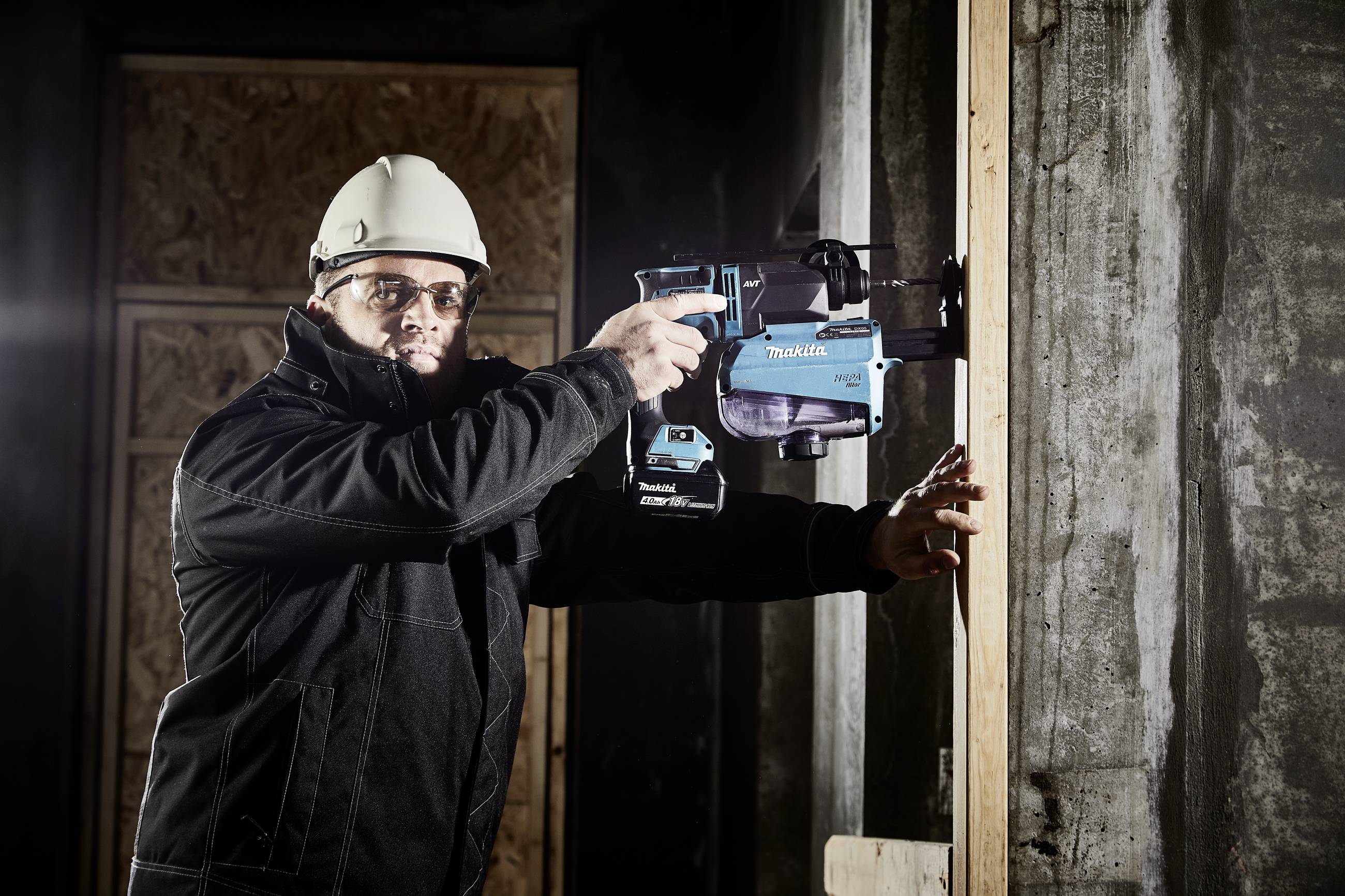A man wearing a hard hat and safety glasses is drilling into a concrete wall with a blue drill. Construction site scene with wooden frames in the background.