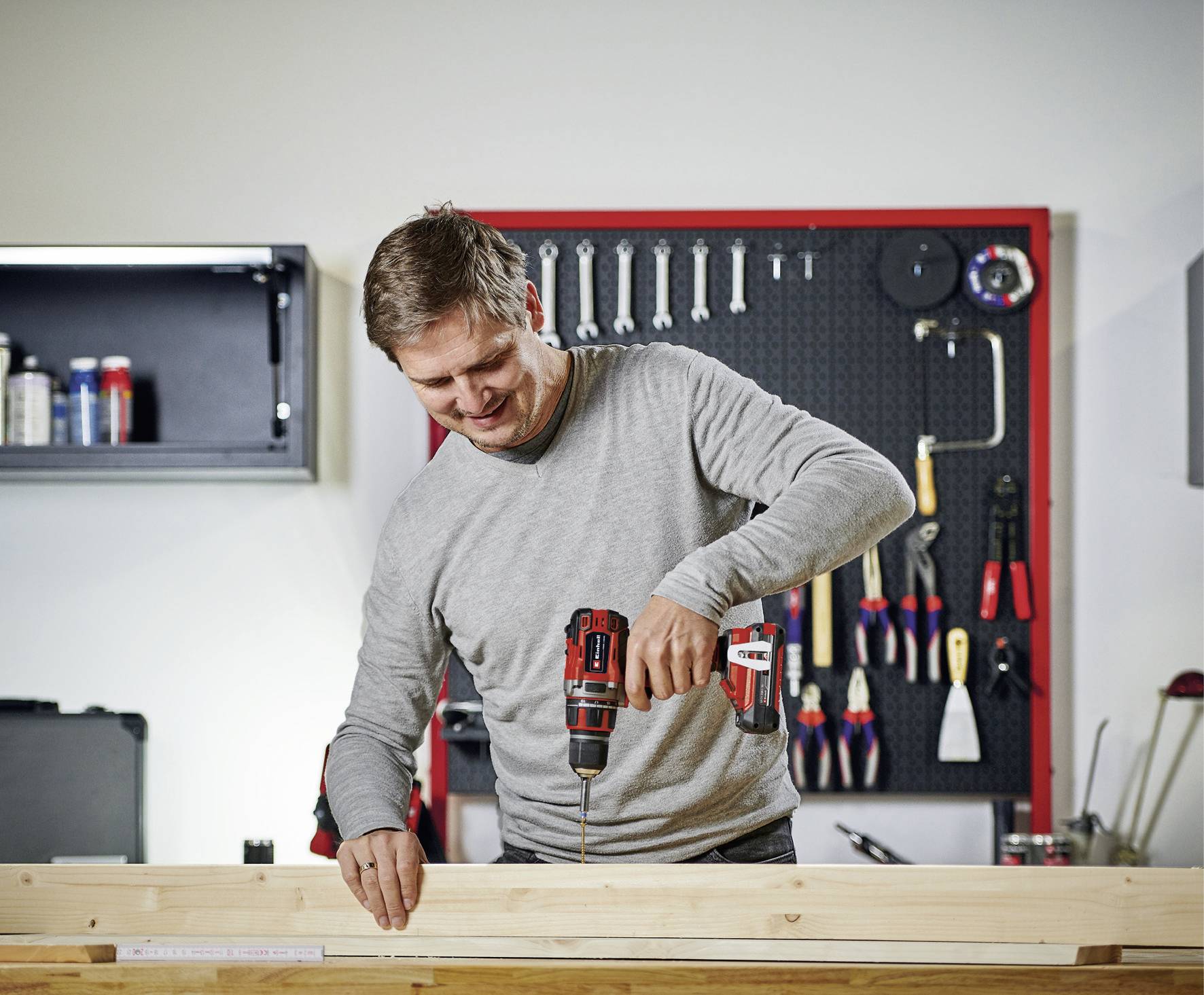 A man is drilling into a wooden board with a cordless drill. Tools are hanging on a wall in the background.