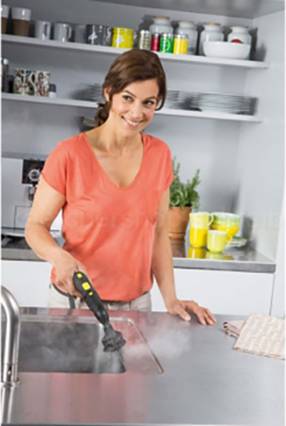 A woman is cleaning the sink with a steam cleaner in a modern kitchen, laughing. Kitchen cabinets with crockery are visible in the background.