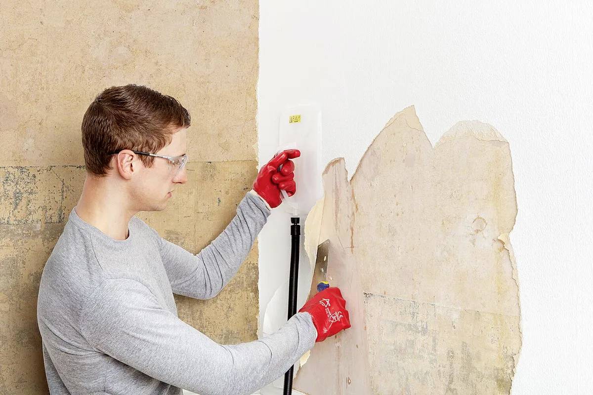 A person is removing old wallpaper from a wall using a scraper and steam device, wearing safety goggles and red gloves.