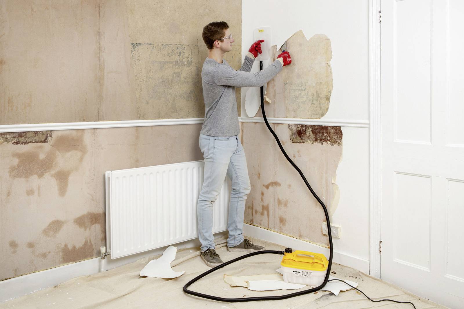 Man removes wallpaper from the wall using a steam device in a room in need of renovation.