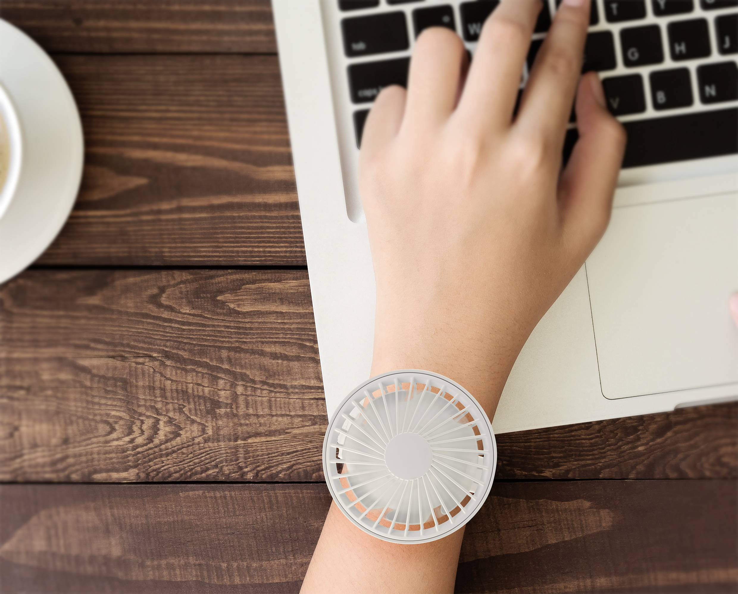 A hand types on a laptop keyboard; a small, round fan is attached to the wrist.