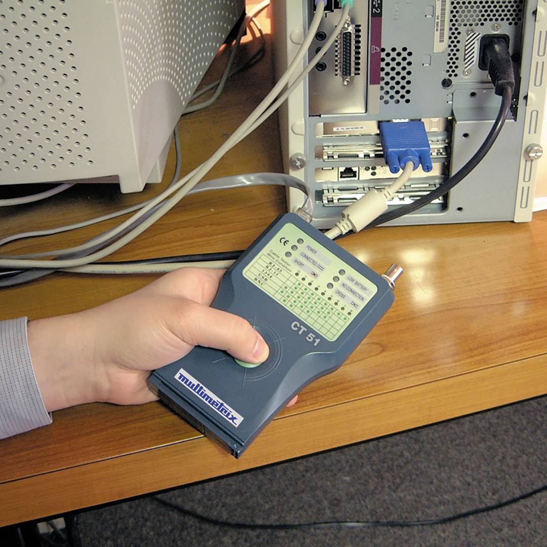 A hand is holding a cable testing device connected to a computer tower. The device displays the label 'multimètre CT51'.