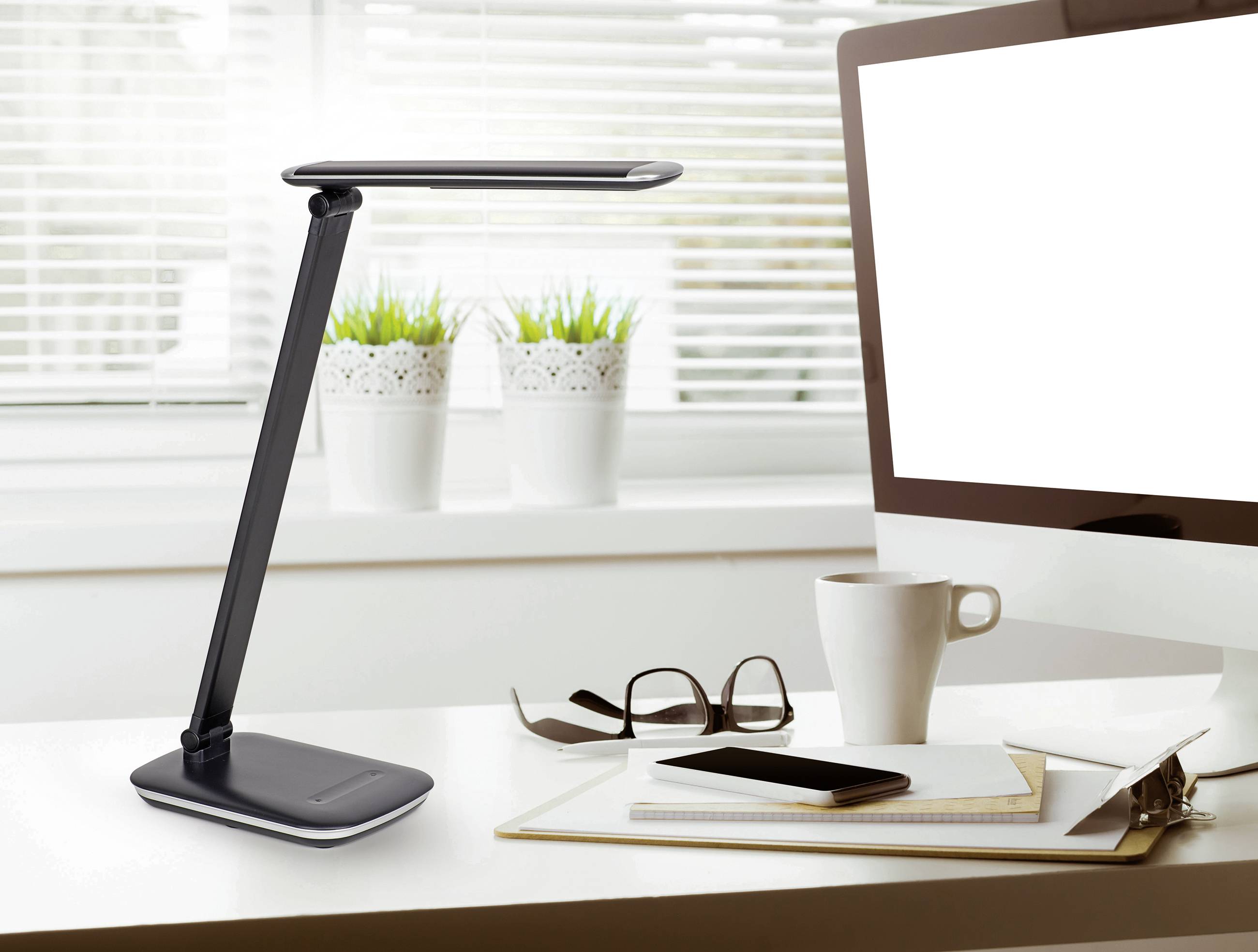 A modern, black desk lamp stands on a desk next to a computer, a pair of glasses, a coffee mug, and notebooks.