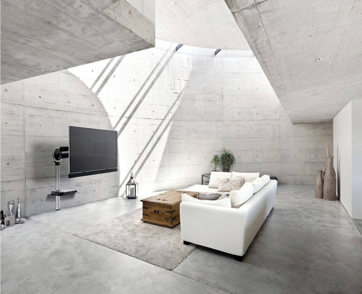 Modern concrete living room with a large sofa, wall-mounted TV and minimalist decor. Plenty of natural light floods in through skylights.