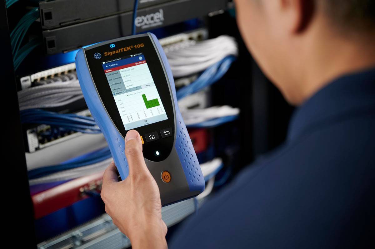 A person is holding a SignaTEK 100 device in front of a network server stack. The display shows a bar graph with network data.