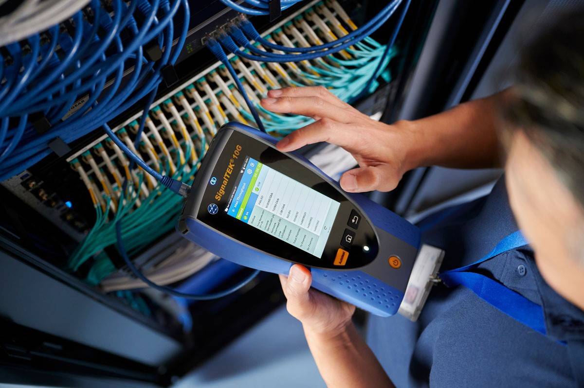 A person is operating a network diagnostic device in front of a server rack with numerous cables to check connections and signal strength.