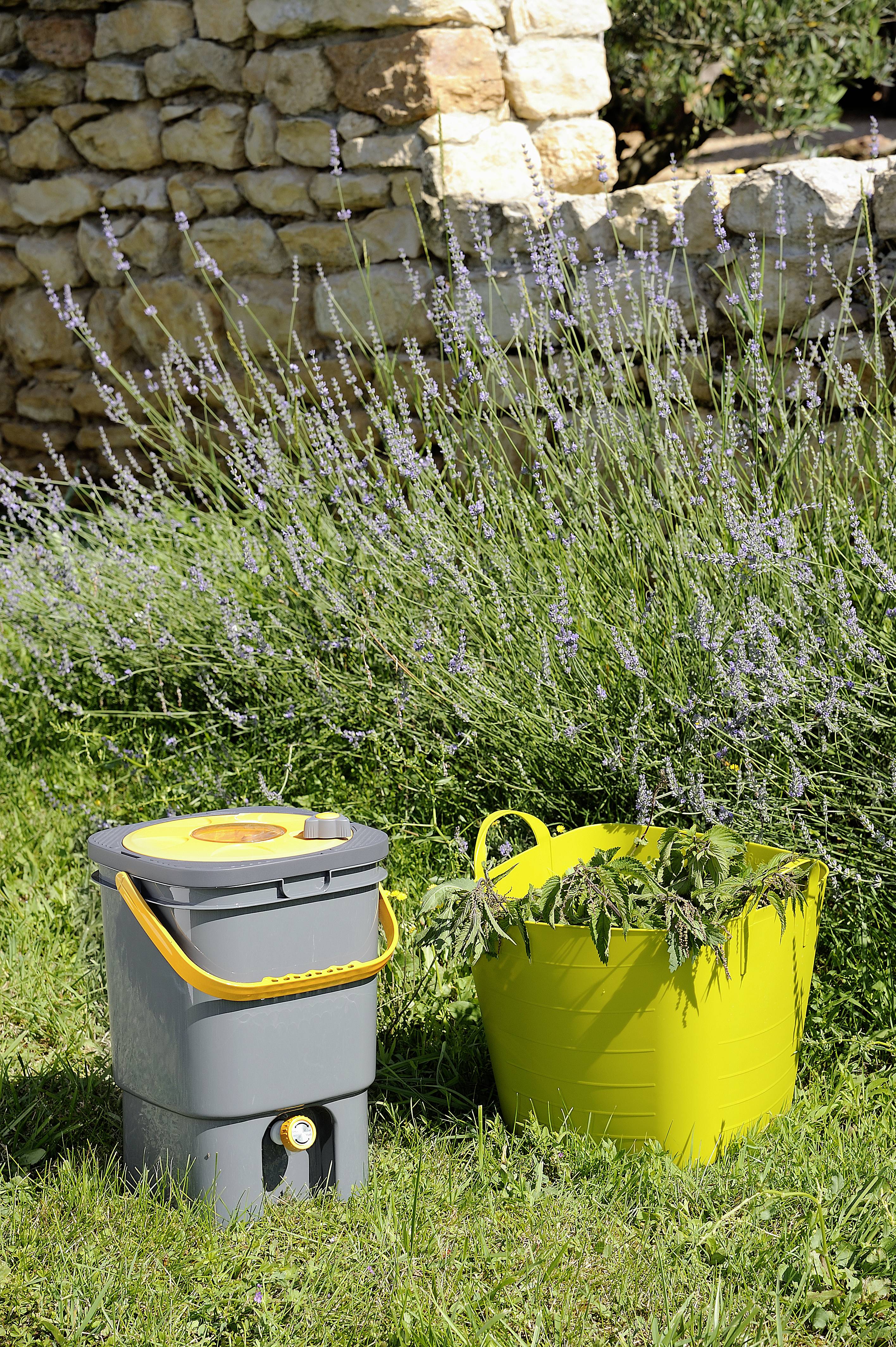 Grey compost bin and yellow bucket with plant waste in a garden in front of a lavender bush. Background: Stone wall.