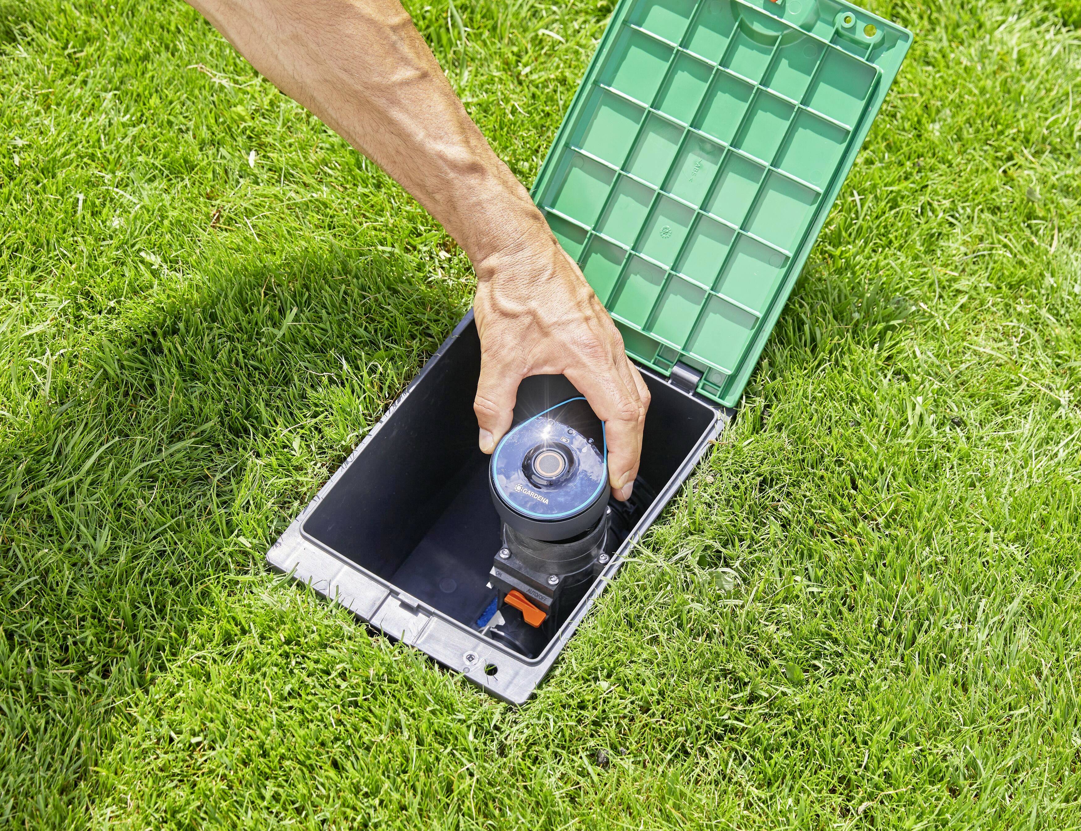 A hand is installing an automatic irrigation sensor in a green lawn valve. The valve's lid is open.