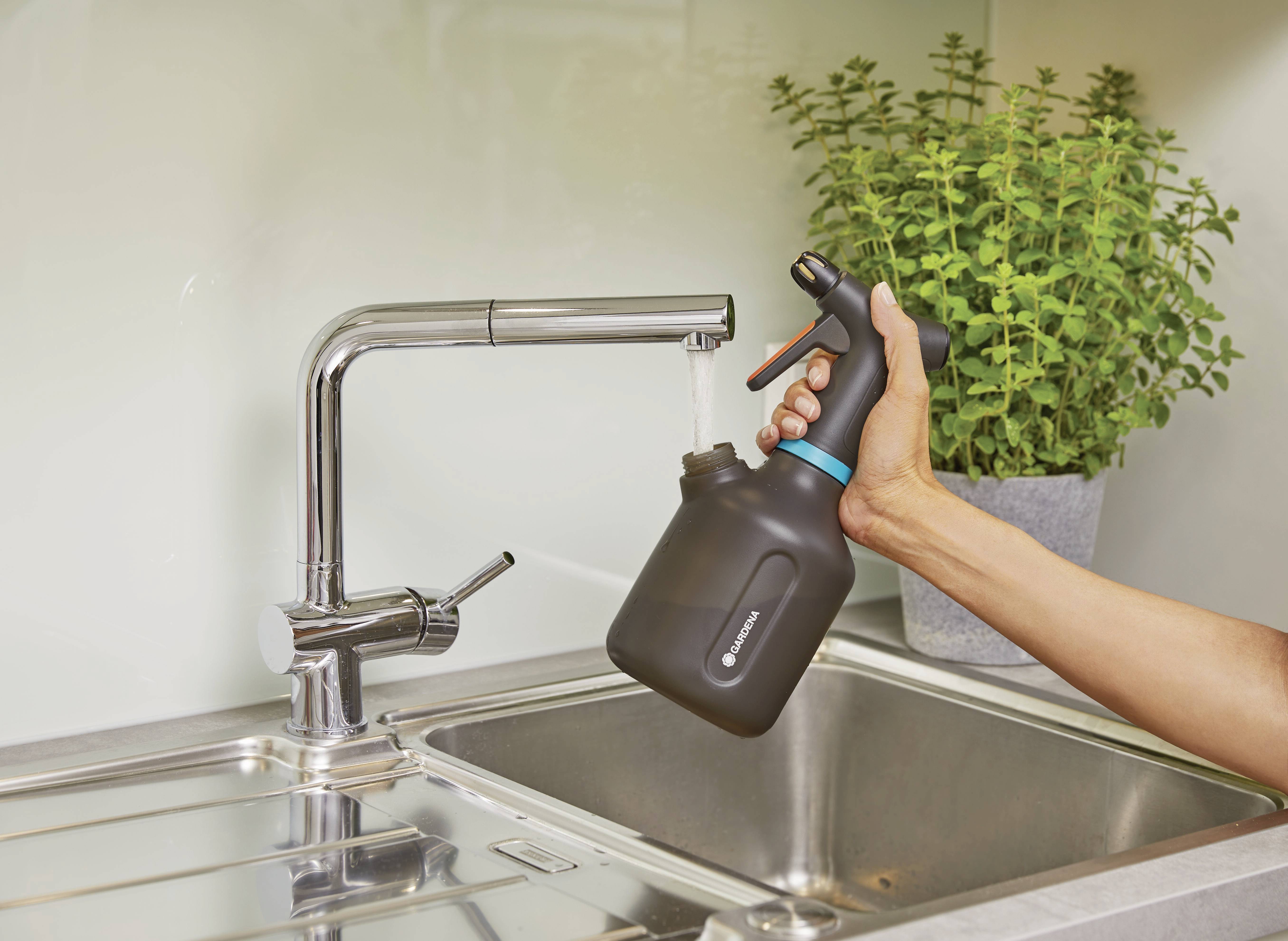 A hand holds a black spray bottle under a tap, from which water is flowing. Next to it stands a pot with a green plant.